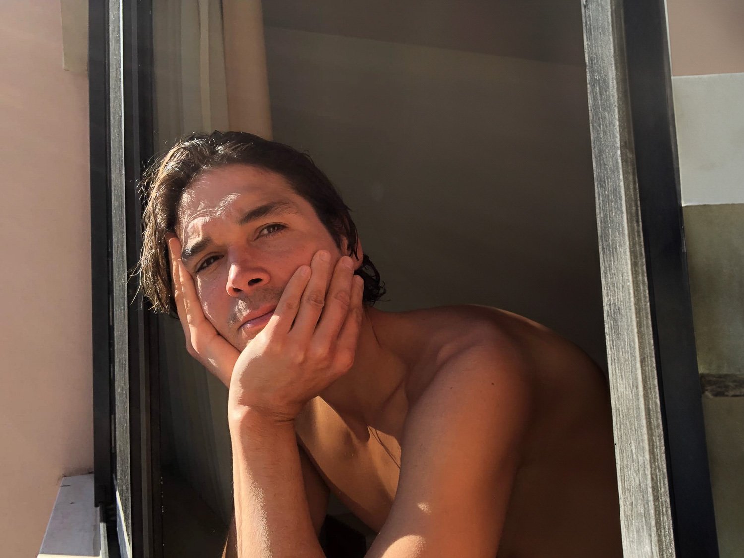A topless man staring out of a sunlit window