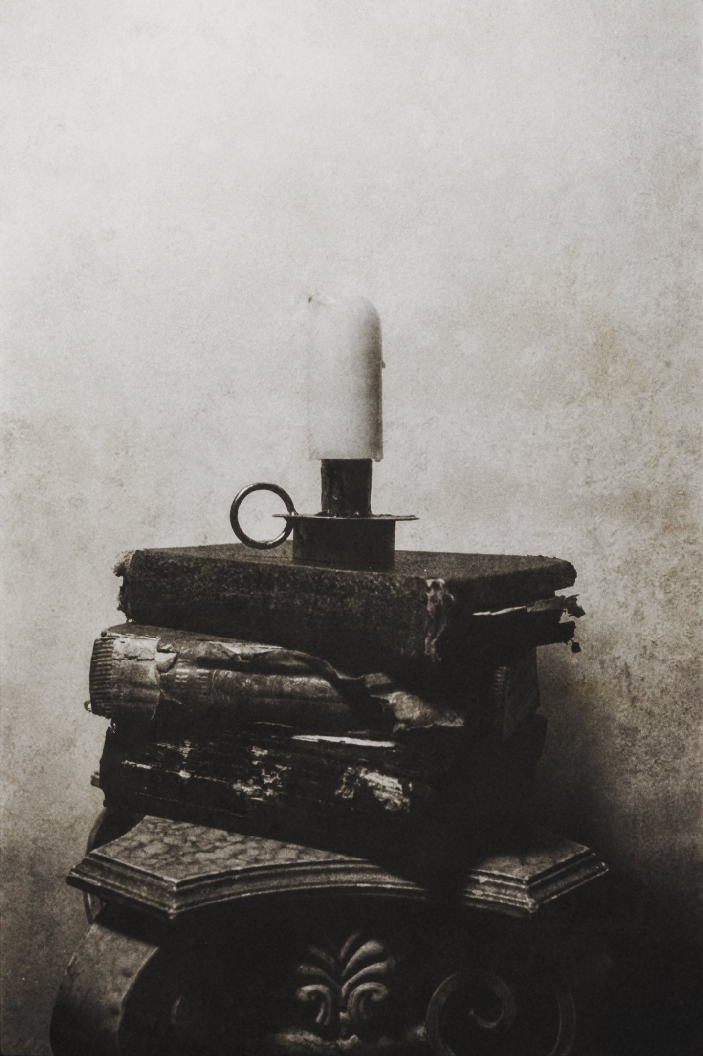 A black and white photograph of a small stack of old books with a candle on top, placed on an ornate, curved table.