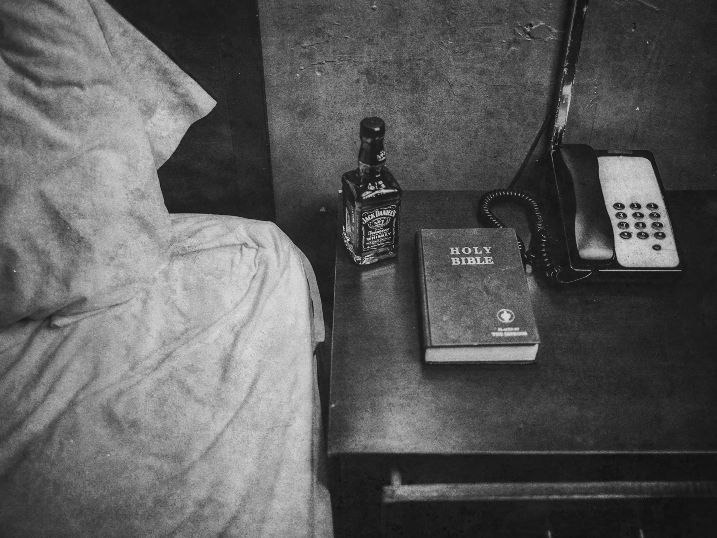 Side of a bed with a nightstand next to it. The nightstand has a bottle of Jack Daniel's whiskey, a Holy Bible, and a landline telephone.