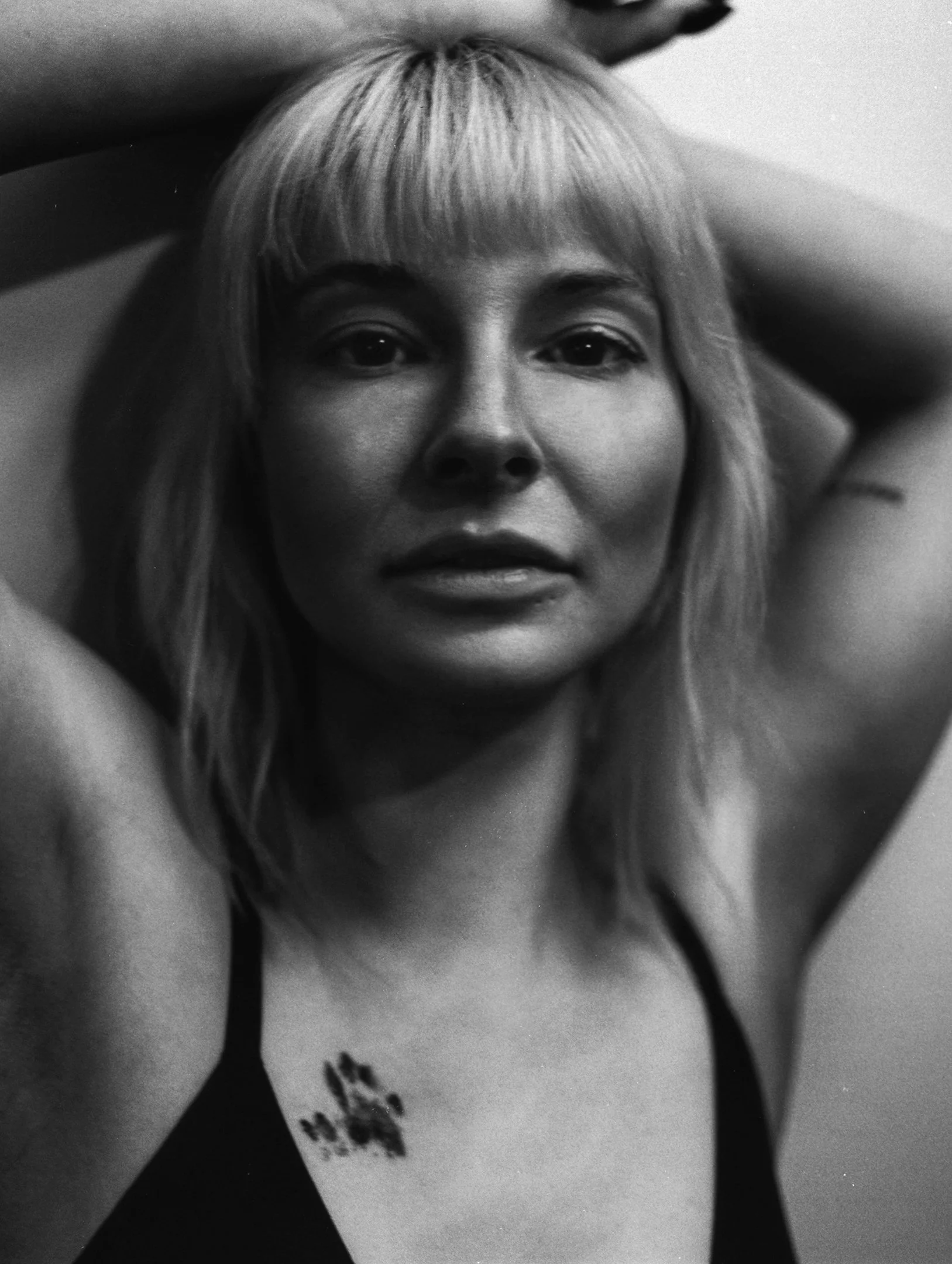 A black and white photo of a woman with light-colored hair, looking directly at the camera with one arm raised and her hand resting on her head.