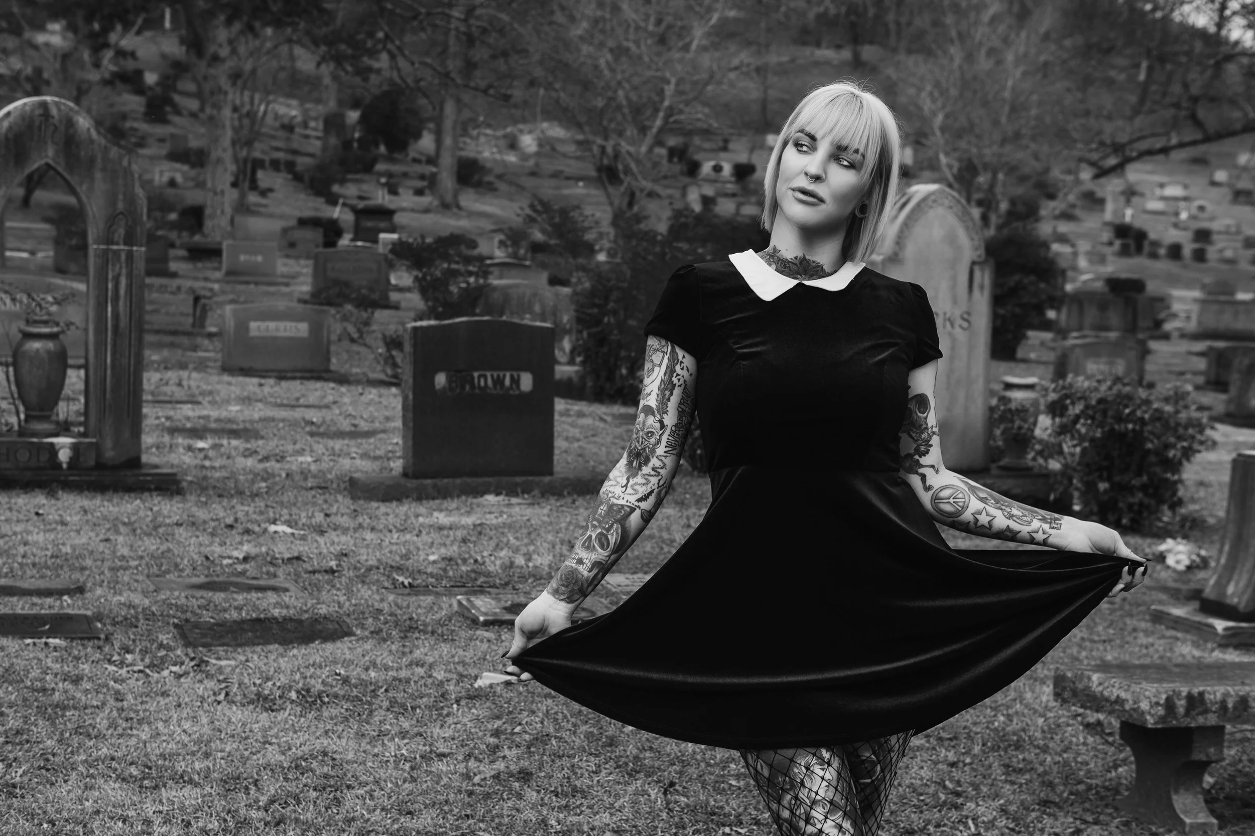 A woman with platinum blonde hair, tattoos on her arms, wearing a black dress with a white collar, standing in a graveyard with tombstones and trees in the background.