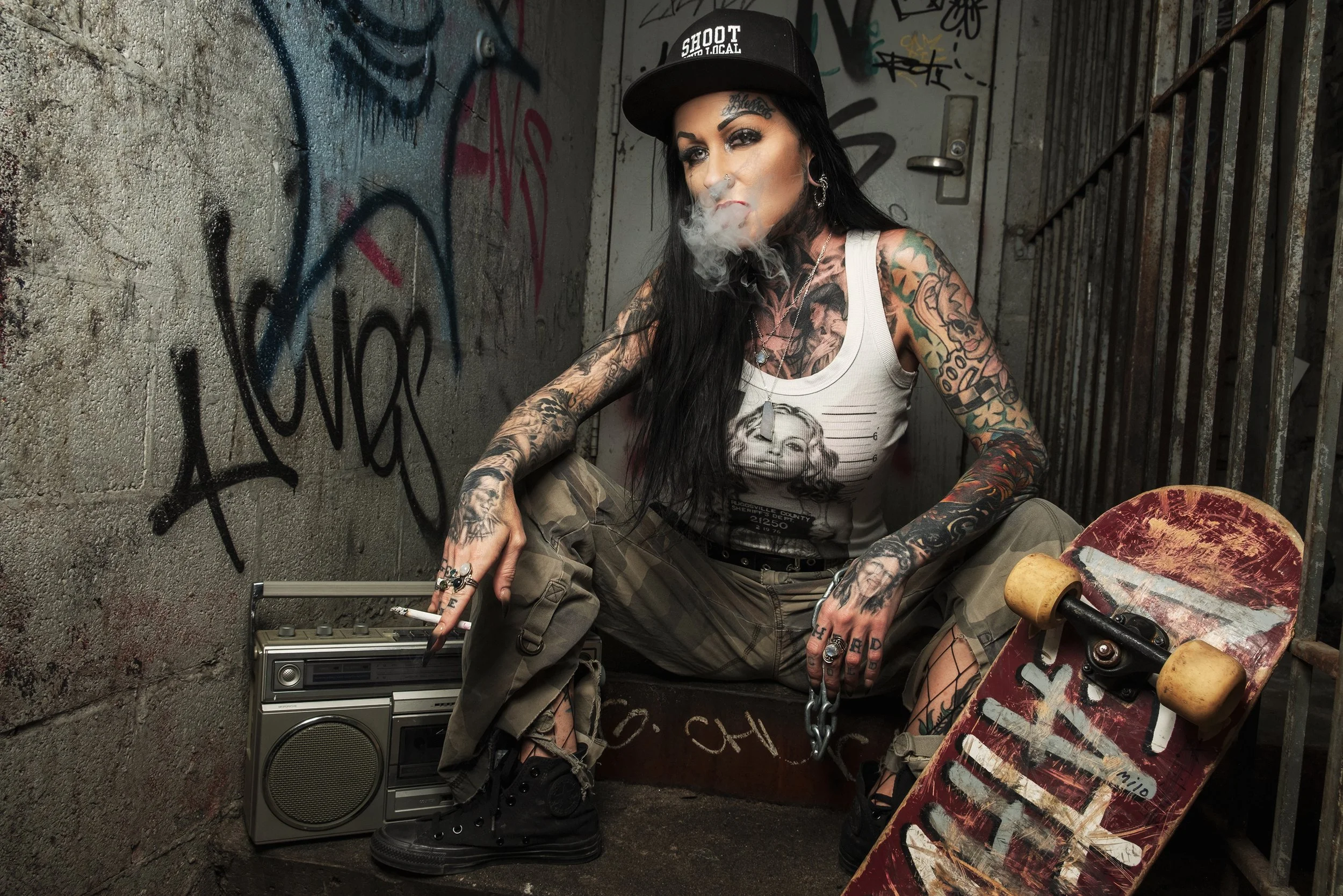 A tattooed woman with black hair wearing a black cap, white tank top, and camouflage pants sitting in an urban alleyway. She is smoking and has a skateboard, an old radio, and a boombox nearby, with graffiti-covered walls in the background.