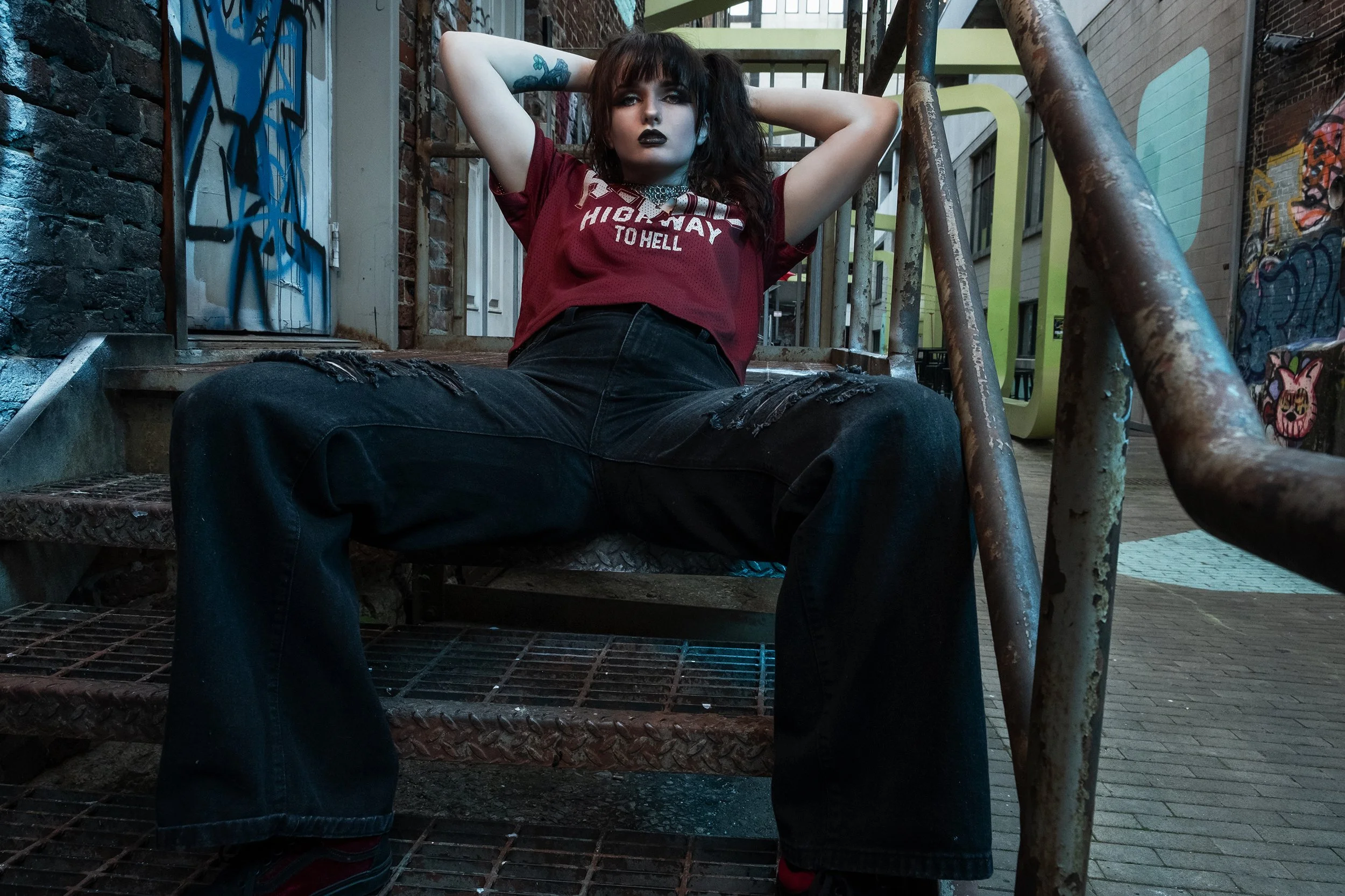 A woman with dark hair, dark makeup, and tattooed arms sitting on metal stairs in an urban alley surrounded by graffiti and brick walls. She wears a red sports jersey, ripped black jeans, and has her hands behind her head.