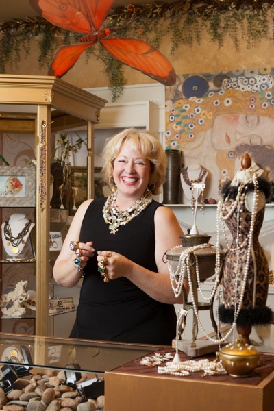 From Erin MacGeraghty’s imagination comes fanciful rings, necklaces and earrings by Linda Hubbard Gulker on December 9, 2013 Editor’s note: With the holiday gift buying season in full swing, we’re visiting some of Menlo Park’s locally-owned stores in the hopes of reminding readers that shopping begins at home. Surrounded by dazzling stones and hovering butterflies, Erin MacGeraghty proudly admits Erin Mac Jewelry in the Sharon Heights Shopping Center in Menlo Park is “not your typical jewelry store.” “I remember one time I was sitting on the floor with my dog,” she recalled. “A woman walked in and sat on the floor next to me. The result was a new pendant for her.” Erin started working at what had been her parents’ store as a teenager and has been operating it on her own since 1974. A transforming moment was the birth of her son 15 years ago. “He’s always been a magical child,” she said. “He grew up here, learned to walk here — hence the double padded carpet. “When he was six, we went to lunch at the Mexican restaurant El Cerrito here in the shopping center and he told me that I should start designing jewelry. I told him that I loved designing for a particular woman and he said, ‘Choose you.’ “So I really started branching out, taking people’s mother-in-law jewelry and transforming it into something they can wear.” As an example of her creativity, Erin showed us her Egyptian pyramid ring (pictured above). “I think and sketch and things pop out,” she said. “The ideas never stop. “I think of them as ‘yes rings.’ Yes you can, yes you deserve it. I really don’t like playing by the rules. That’s why I like designing.” While rings may be her specialty, she also has a wide selection of necklaces and earrings. “Many of the necklaces are Victorian belt buckles I’ve turned into necklaces. The one I’m wearing today I designed in high school.’ Erin views her shop as a place to have fun. “Jewelry is about love,” she said. “For me, it’s about sending a woman off feeling better than when she came in!” Photos by Scott R. Kline Tagged as: Erin MacGeraghty , jewelry stores in Menlo Park