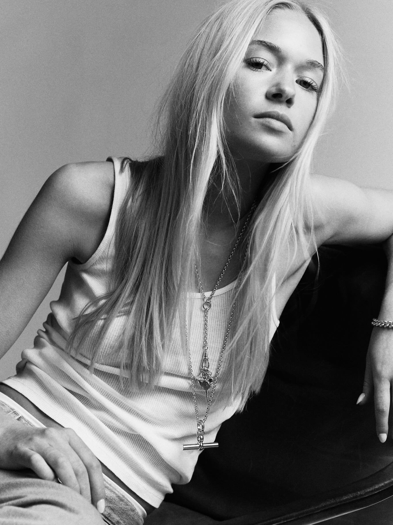 Black and white photograph of a young woman with long blonde hair and light skin, looking at the camera with a neutral expression. She is wearing a sleeveless top with striped pants, layered necklaces, and has her right hand resting on her waist. The background is plain.