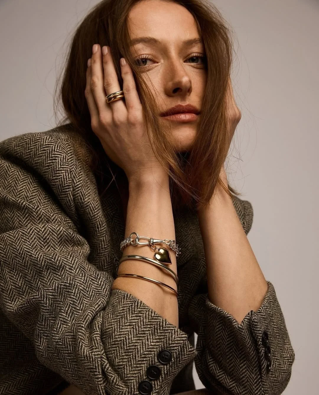 A woman with red hair wearing a brown herringbone jacket, posing with her hands on her face, showcasing multiple bracelets and rings.