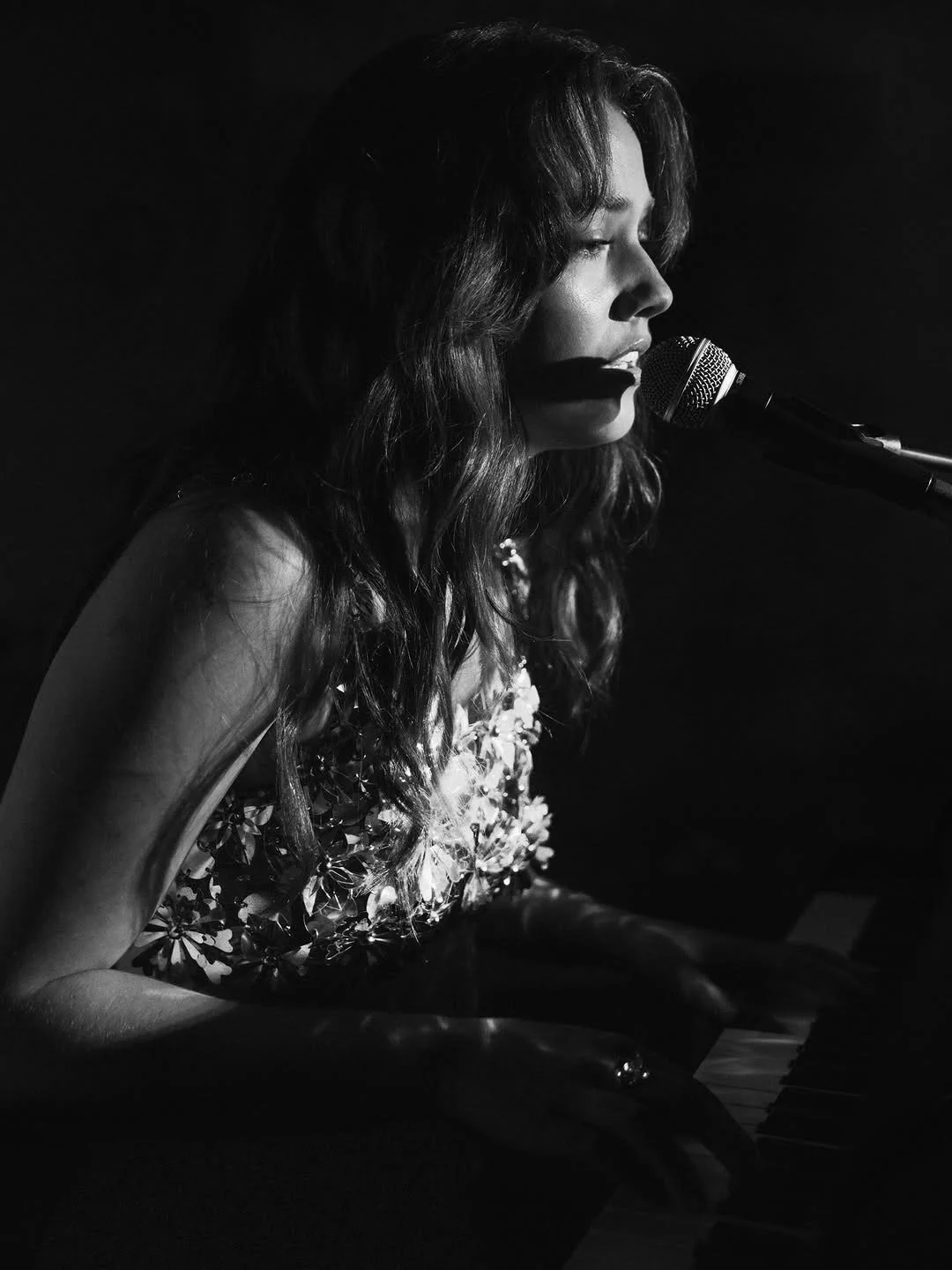 A woman with long wavy hair, seen in profile, singing and playing the piano in a dimly lit setting, with a microphone in front of her.