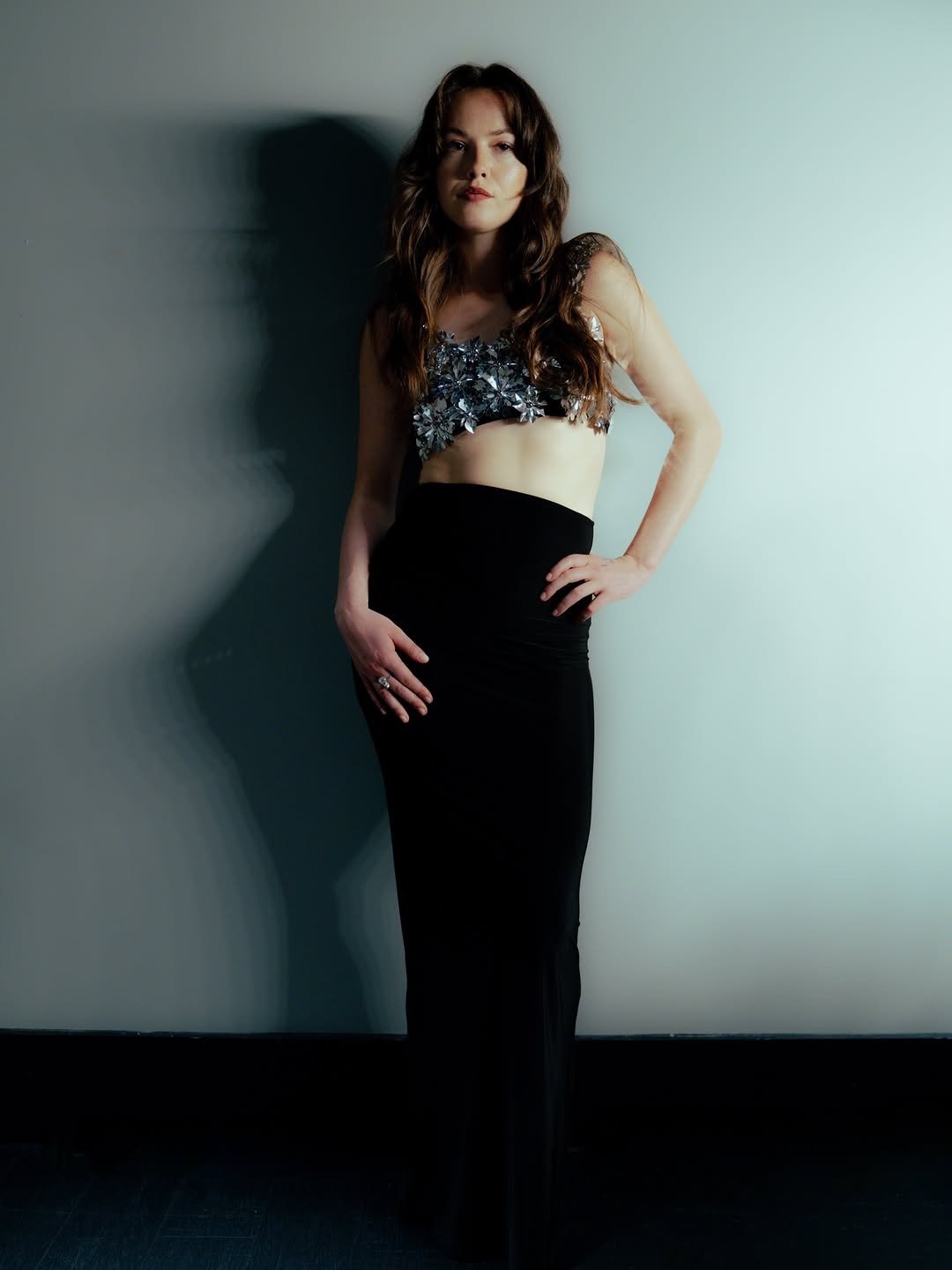 A woman with long brown hair wearing a black skirt and a sparkly crop top posing with her hand on her hip in front of a plain light-colored wall.