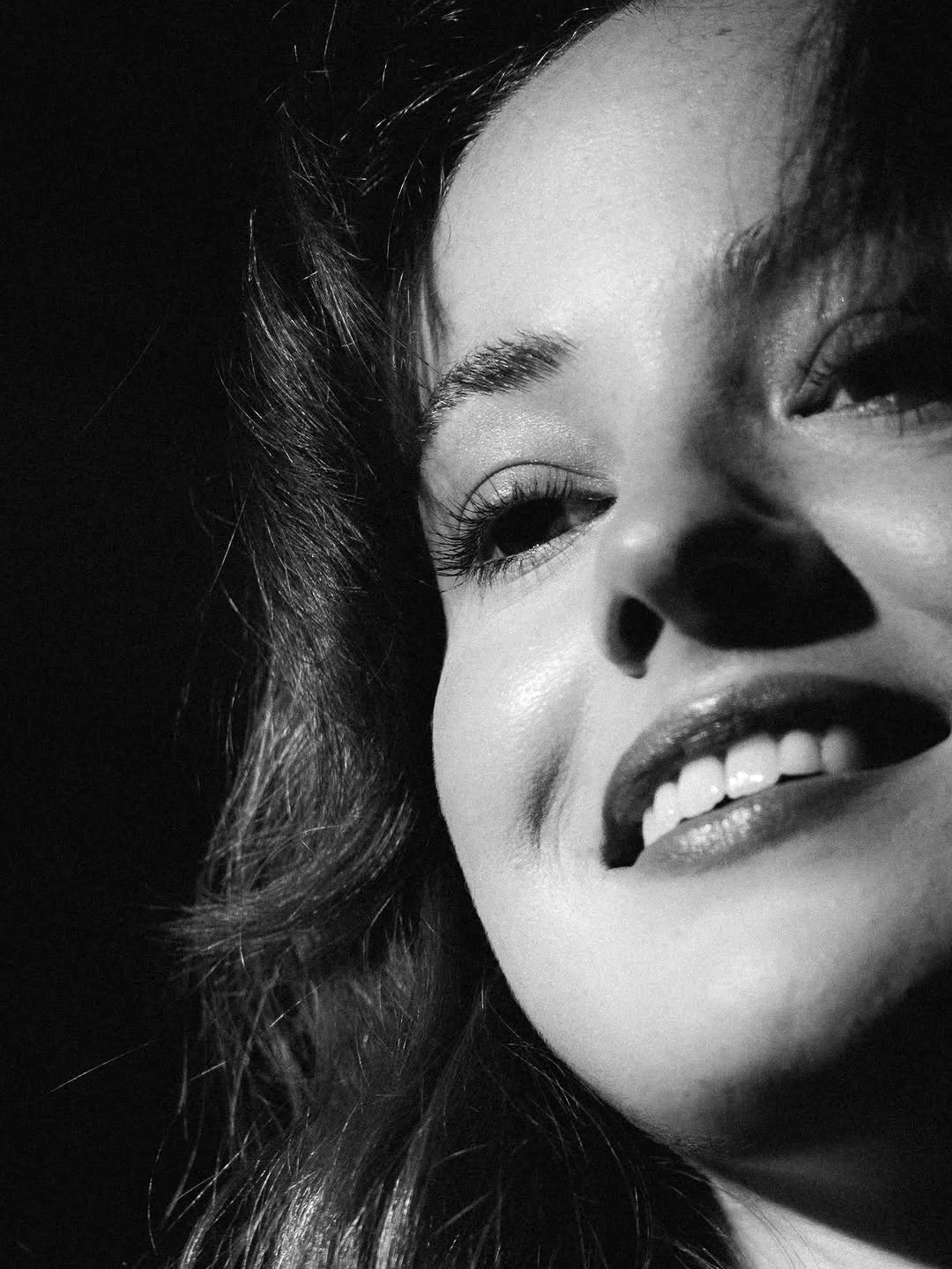 Close-up black and white photo of a woman's face with makeup, smiling with teeth visible, and dark hair.