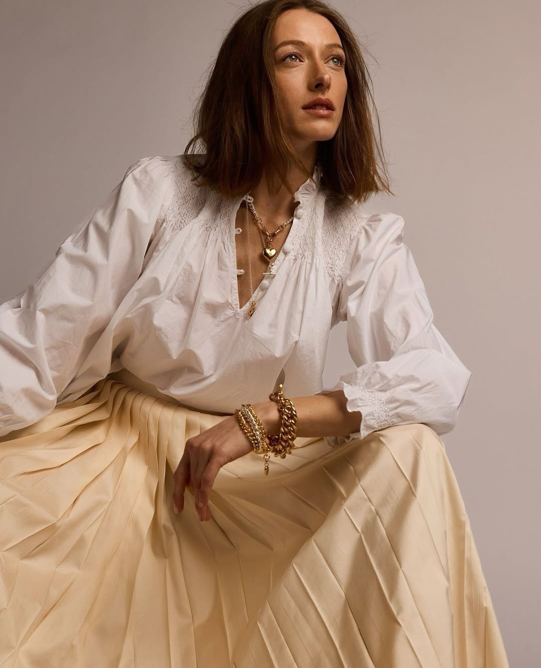 A woman with shoulder-length brown hair wearing a white blouse with lace details and a flowing beige skirt, accessorized with gold jewelry including necklaces, bracelets, and rings, posed against a plain neutral background.