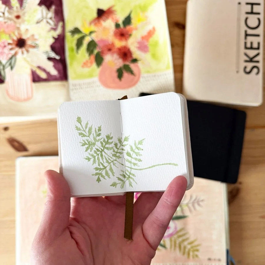 a tiny sketchbook with a fern painting and a pile of sketchbooks filled with flowers on Anne Butera's painting table