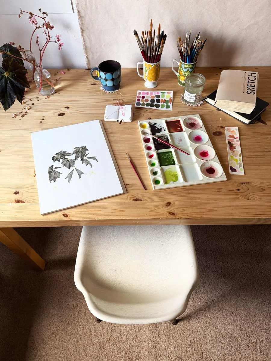 Anne Butera's art table piled with sketchbooks, paint swatches and an in-progress watercolor painting of a begonia plant