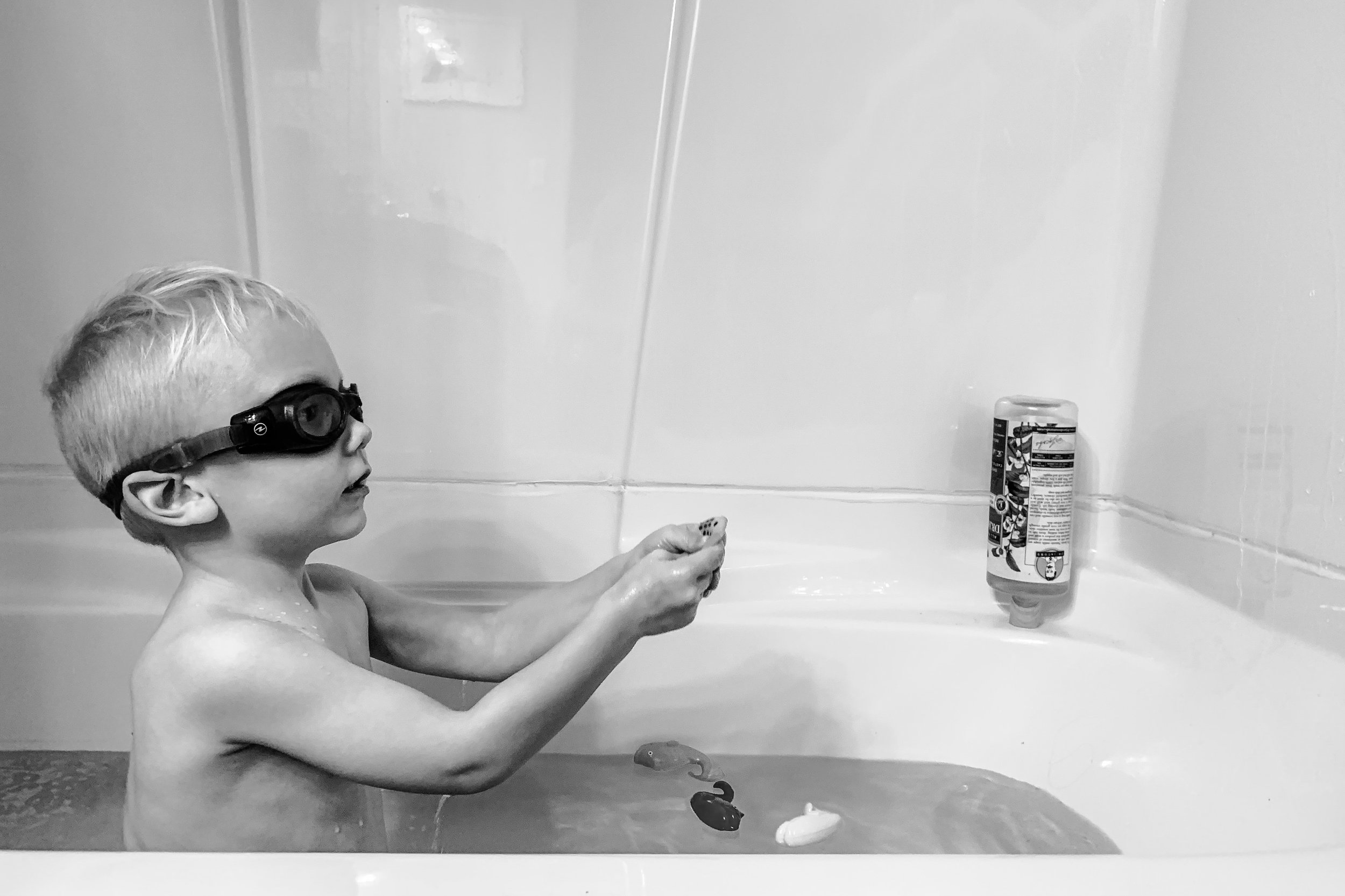 little boy in goggles spraying bath toy at wall of tub
