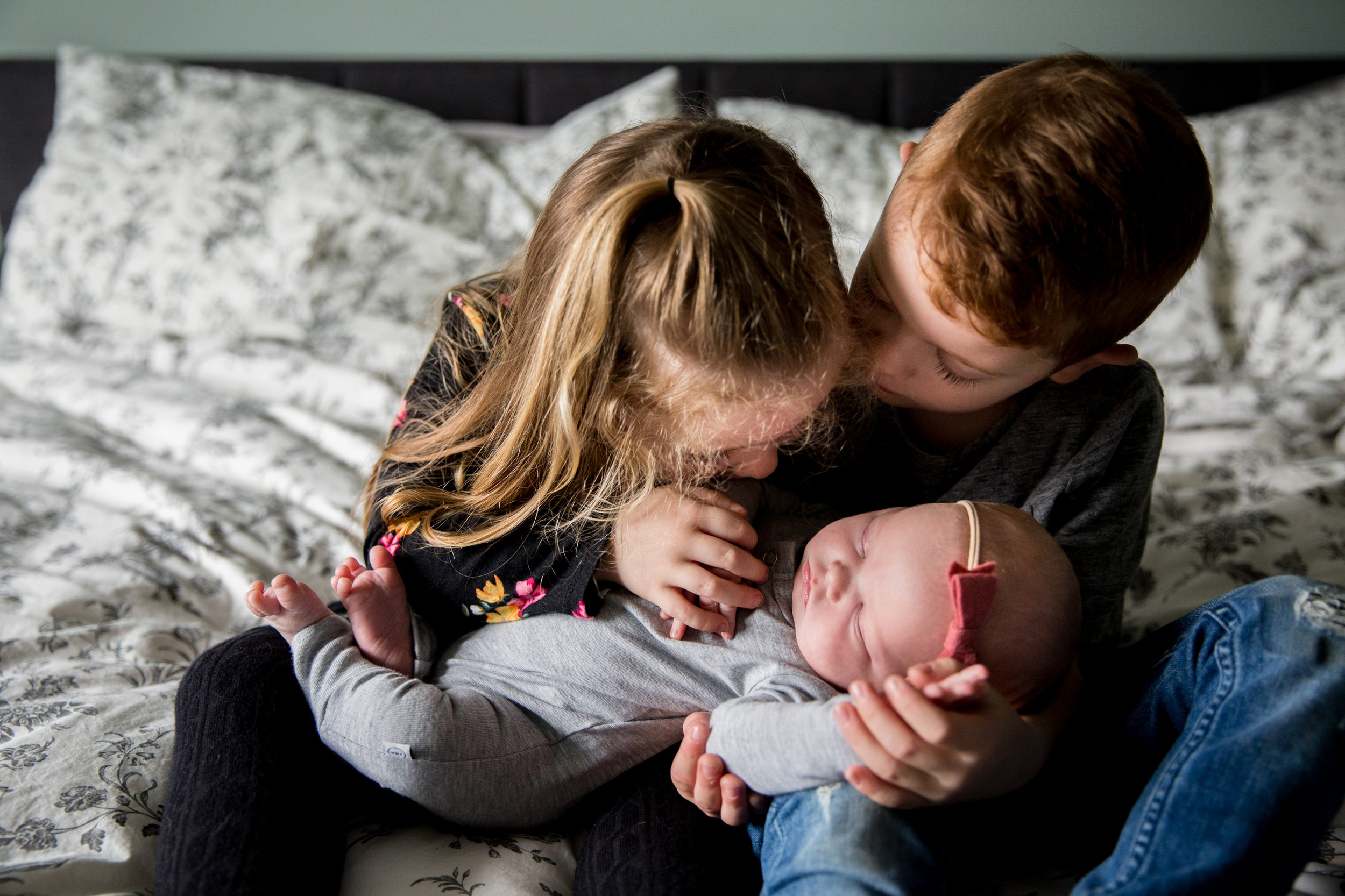 siblings holding baby girl on bed looking at her