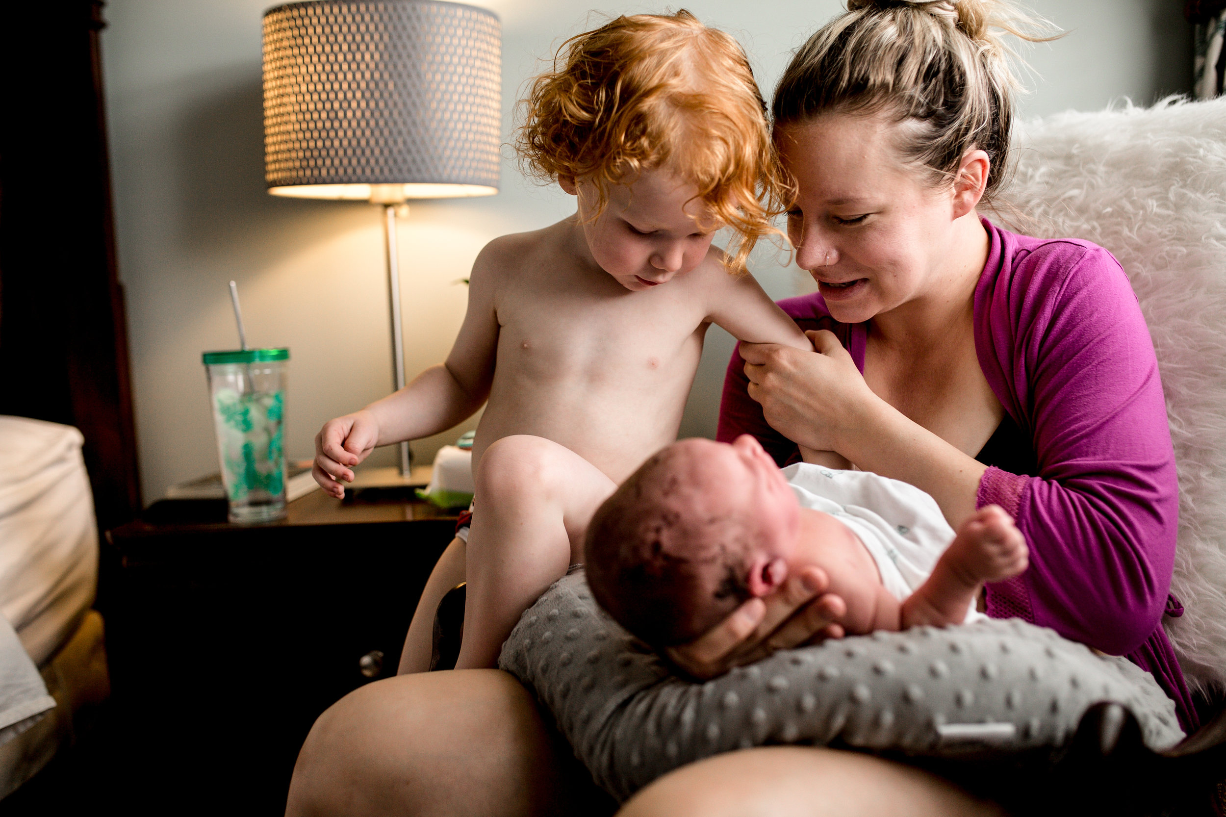 mom sitting on chair in a purple robe helping her toddler look at his new baby brother