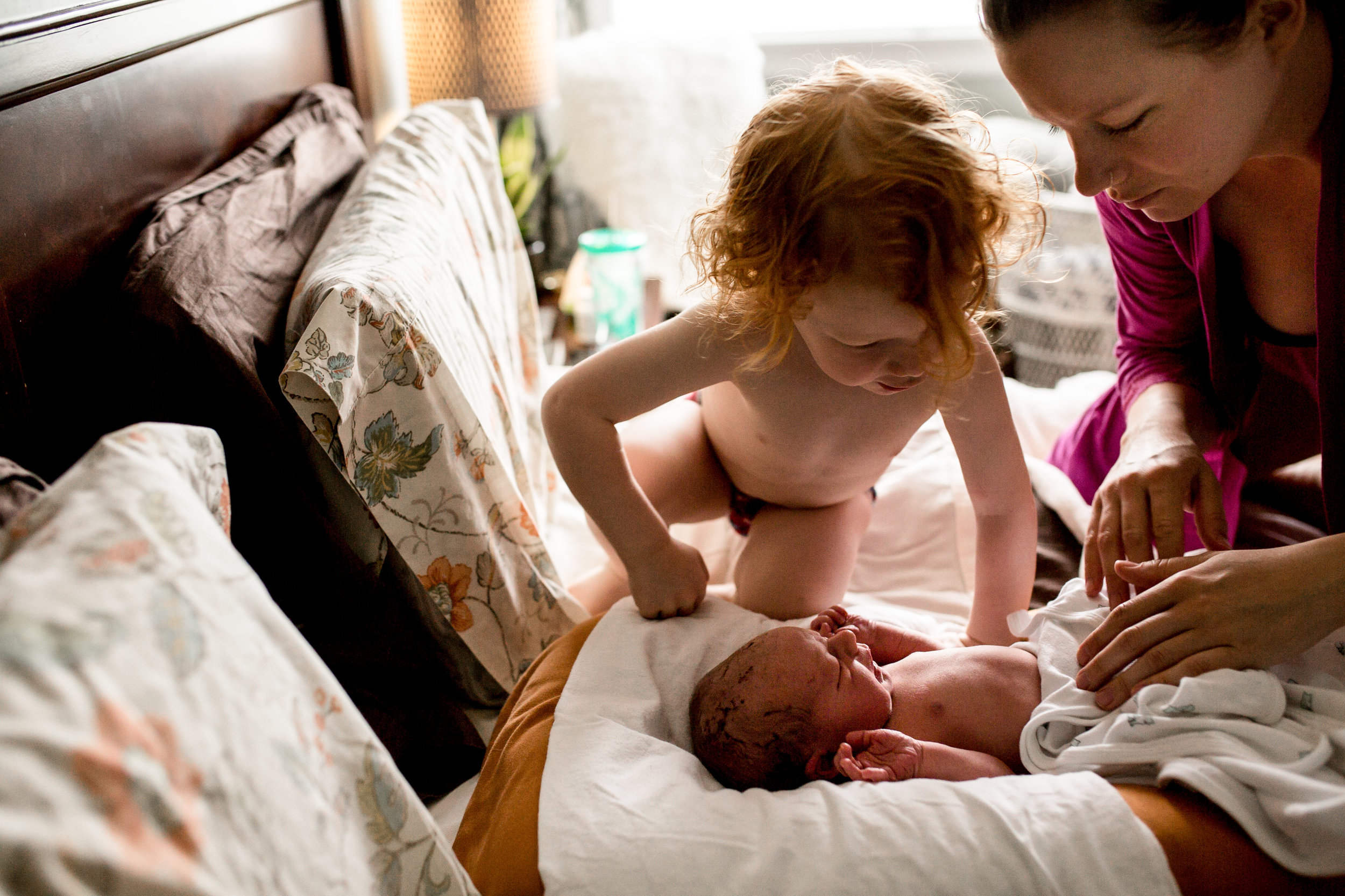 mom and toddler looking down at baby on the bed