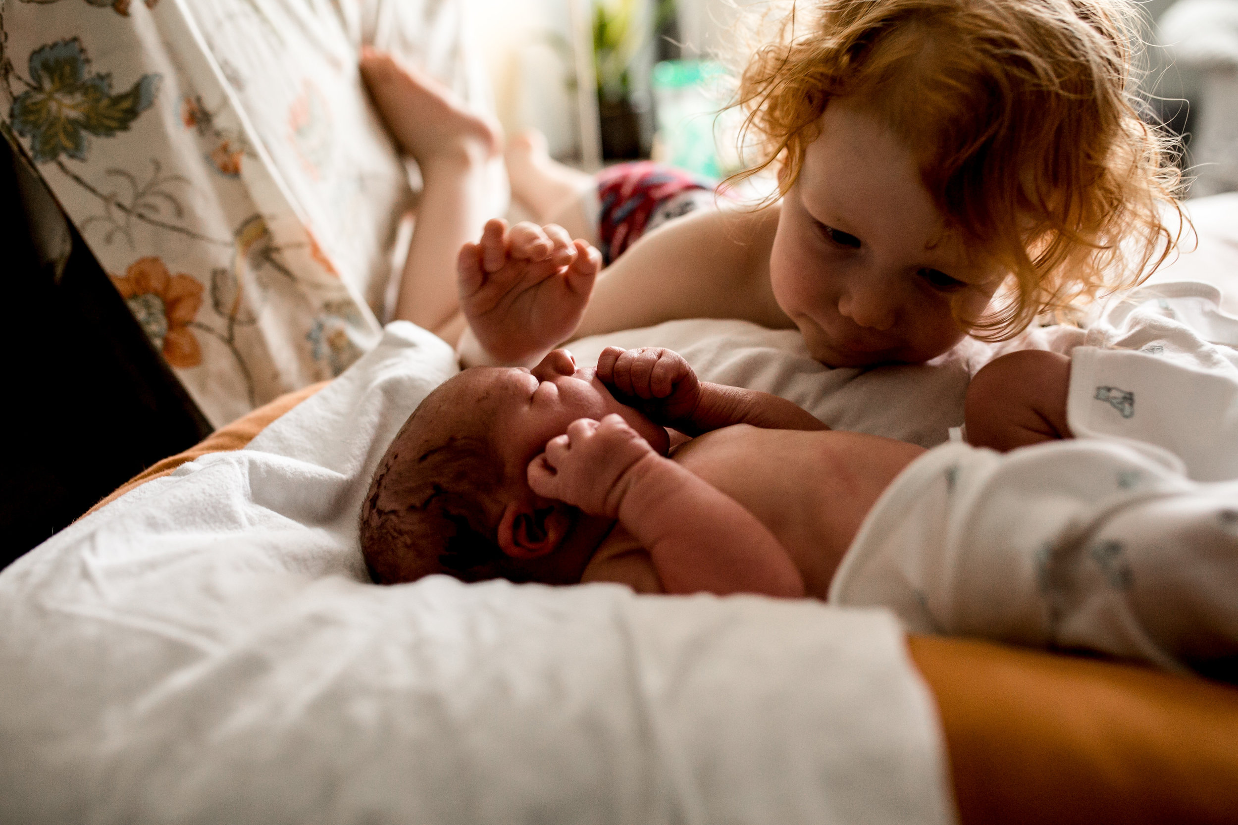toddler boy looking at his baby brother reaching for him on the bed