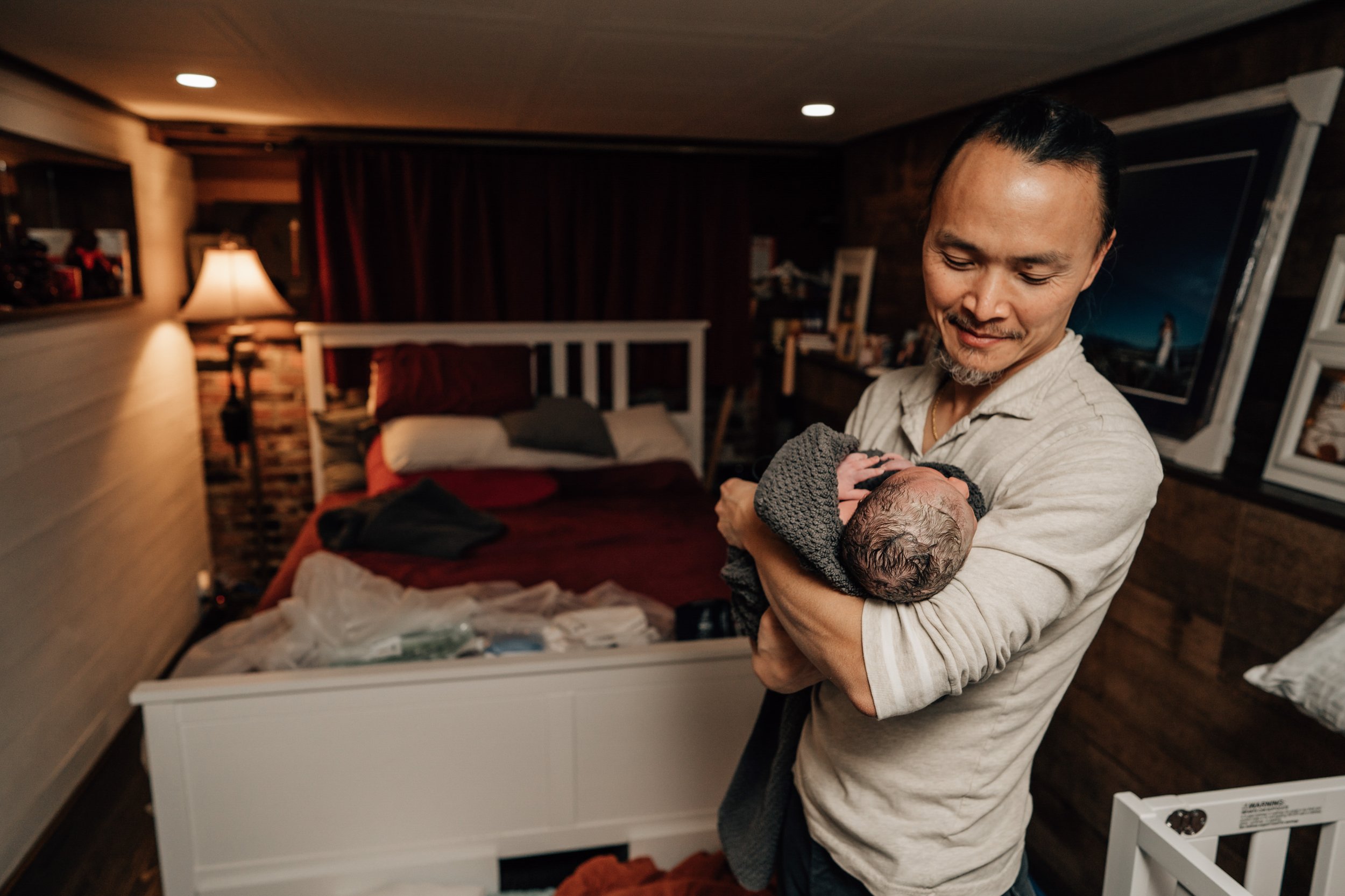 aisian dad holding newborn daughter near calgary inside dark room with lamp.jpg