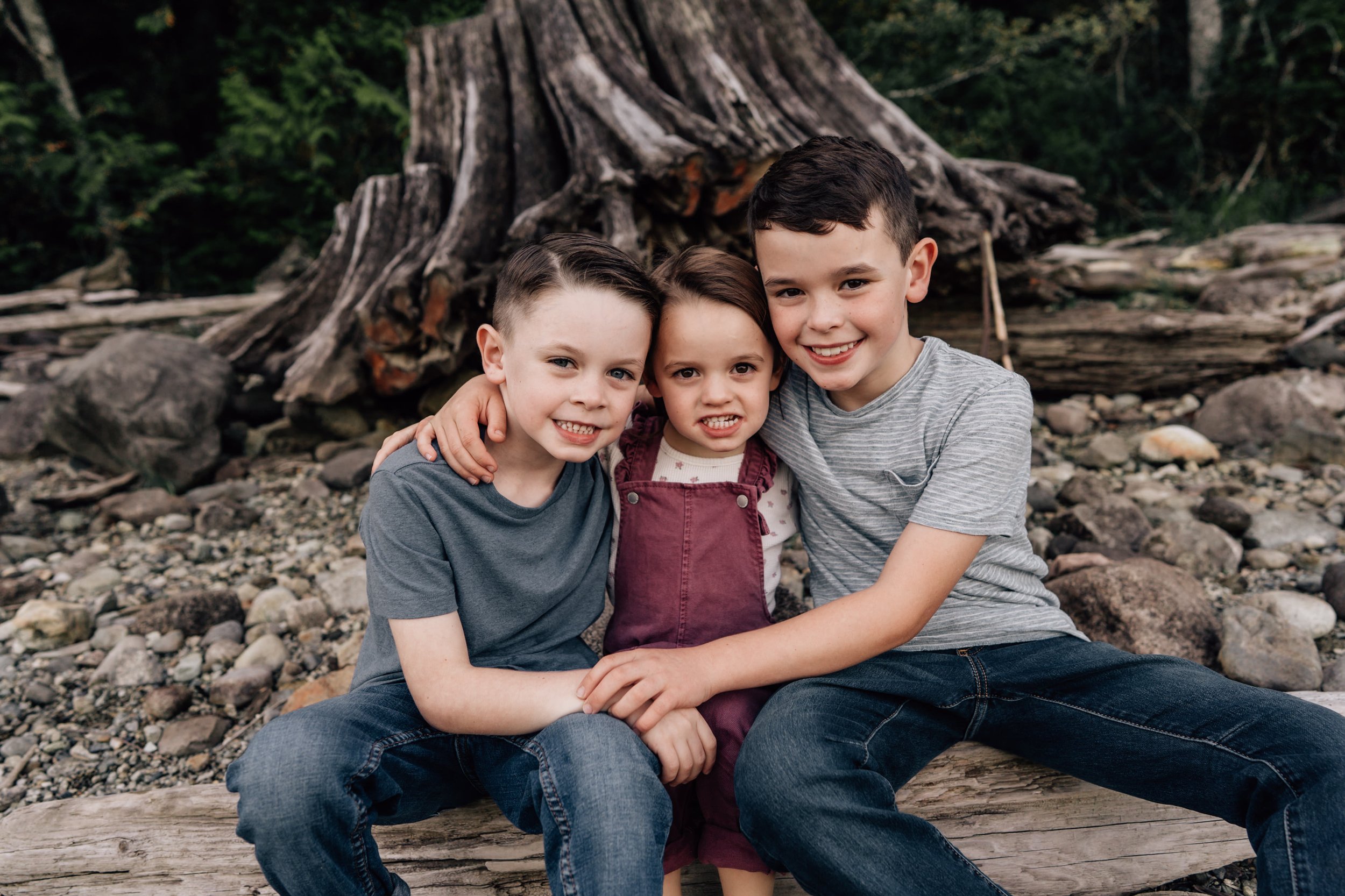 three kids smiling together sitting on a log, two boys and a girl during family photos at rocky beach near calgary