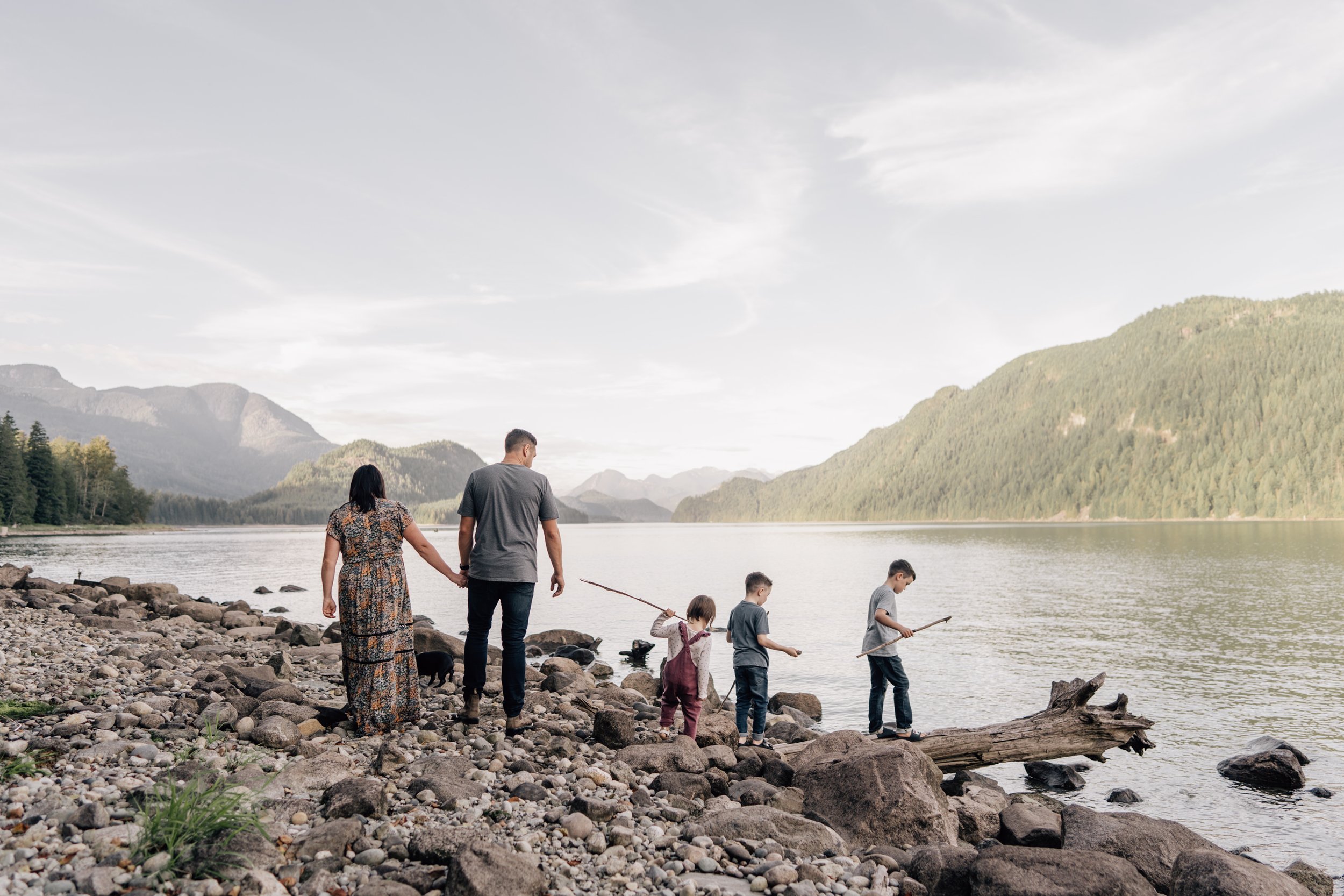 two parents and three kids walking along the shore of a lake with mountains in the background