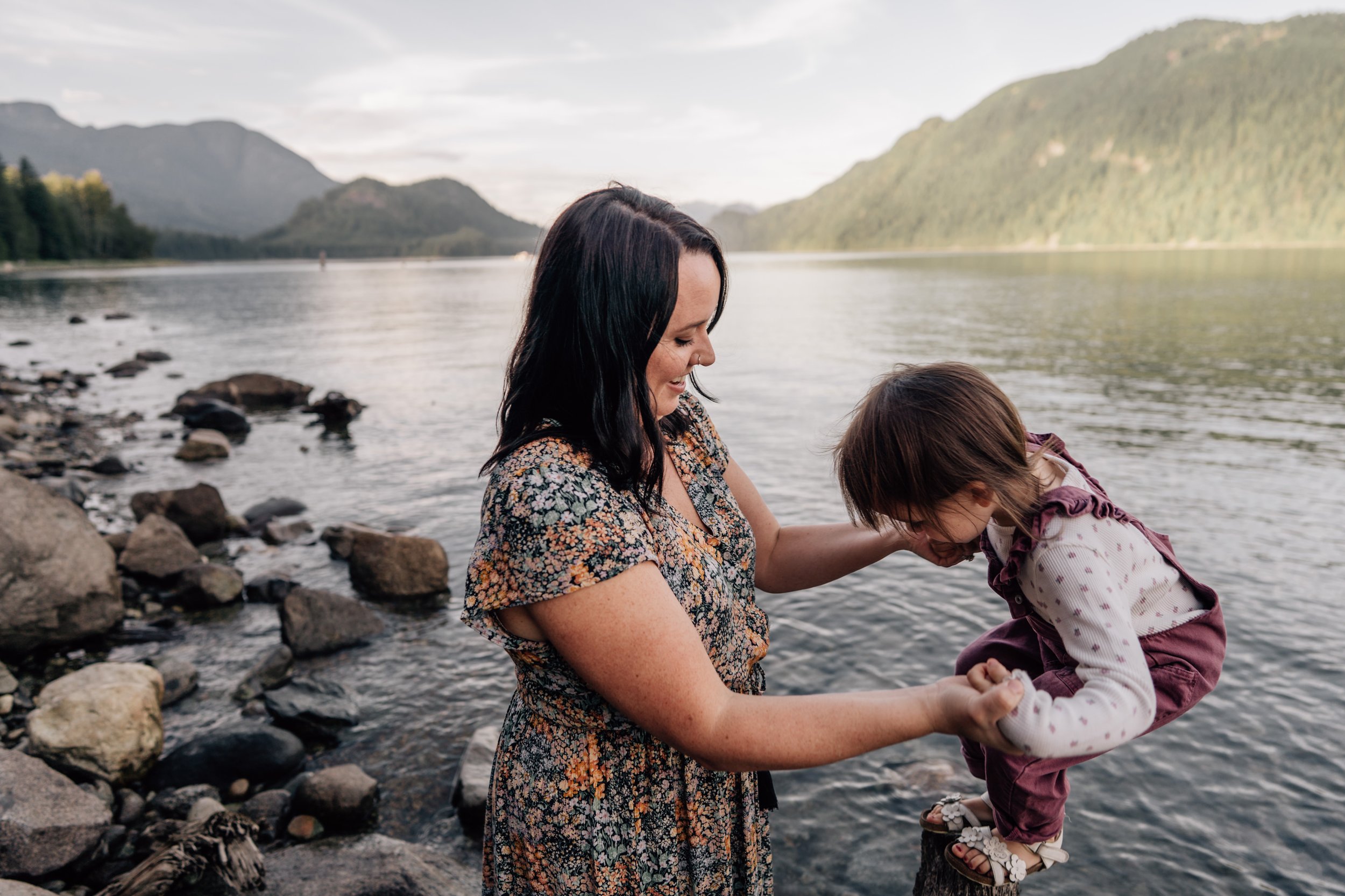 mom holding daughter's hands while she jumps off a log near the shore of the water