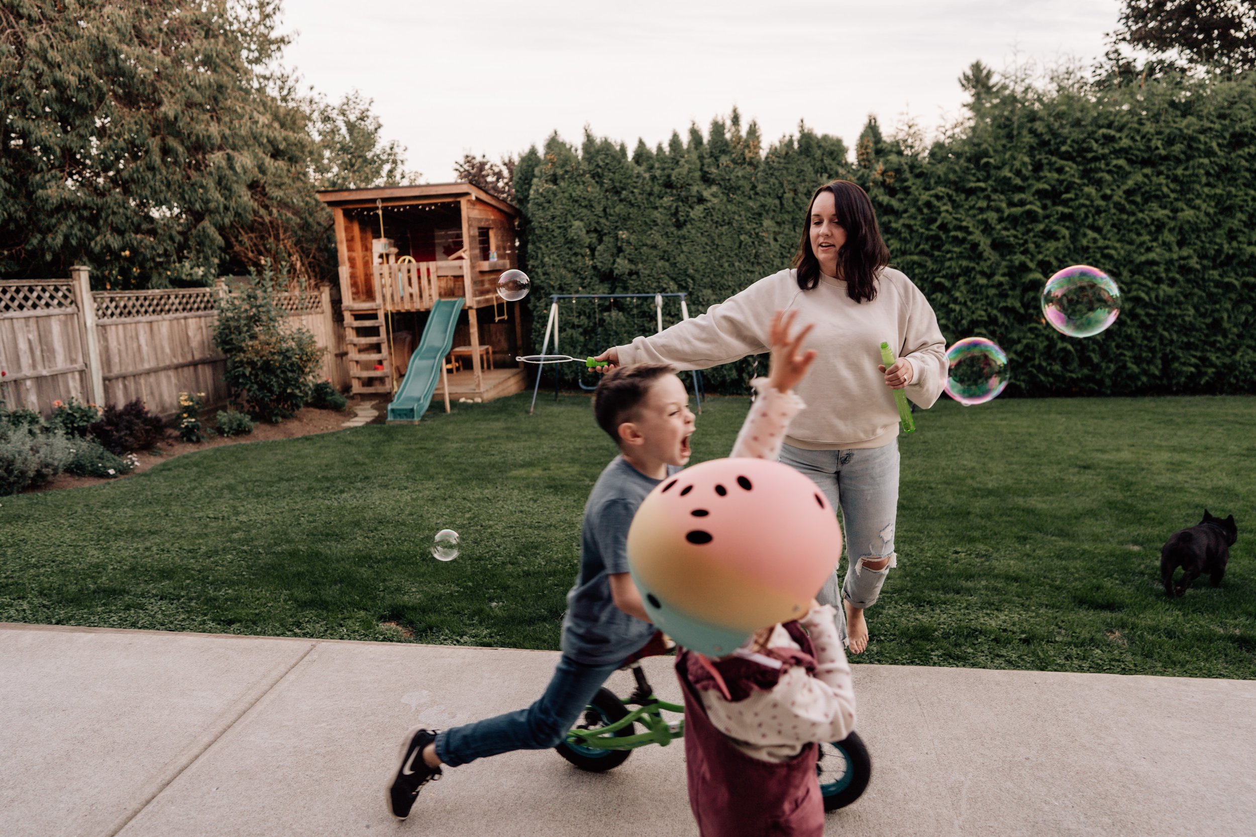 mom using a bubble wand for kids who are running and biking around in the foreground near calgary backyard