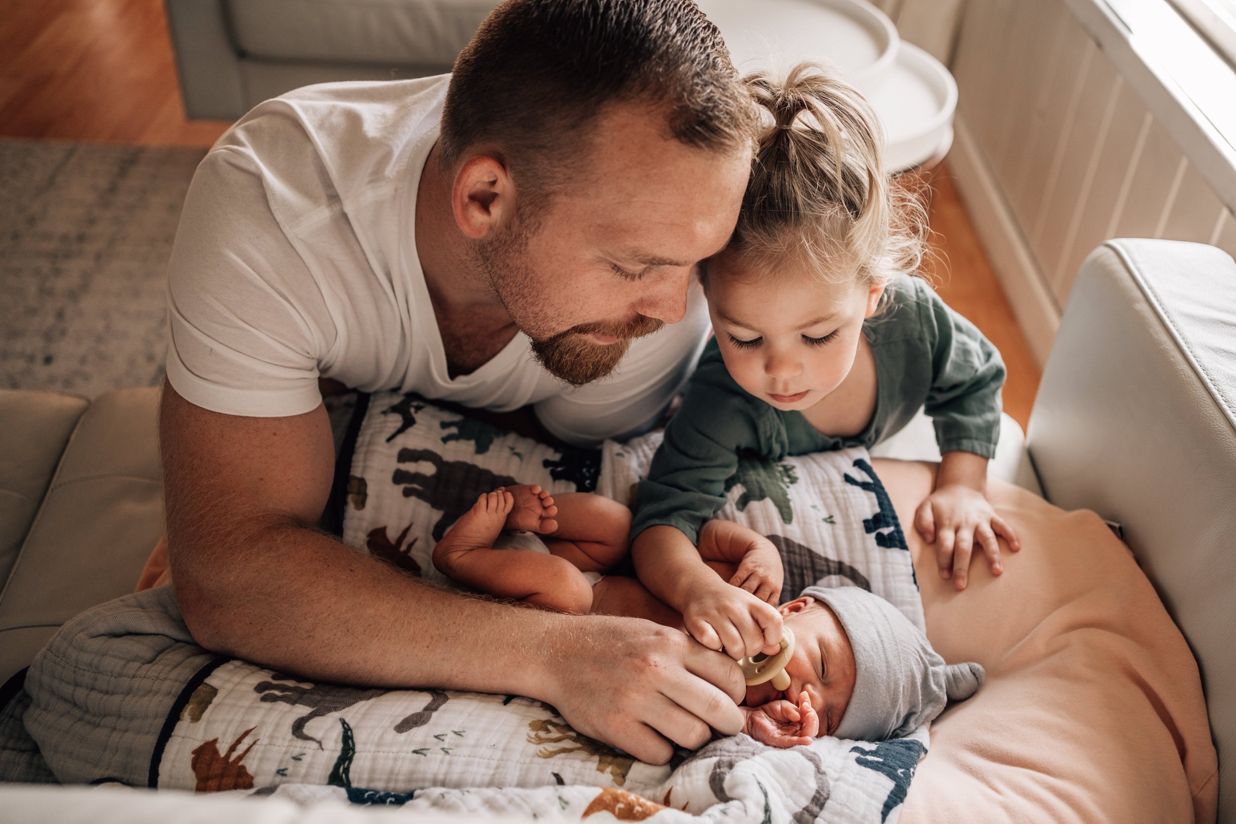 dad and sibling leaning over baby with soother on couch