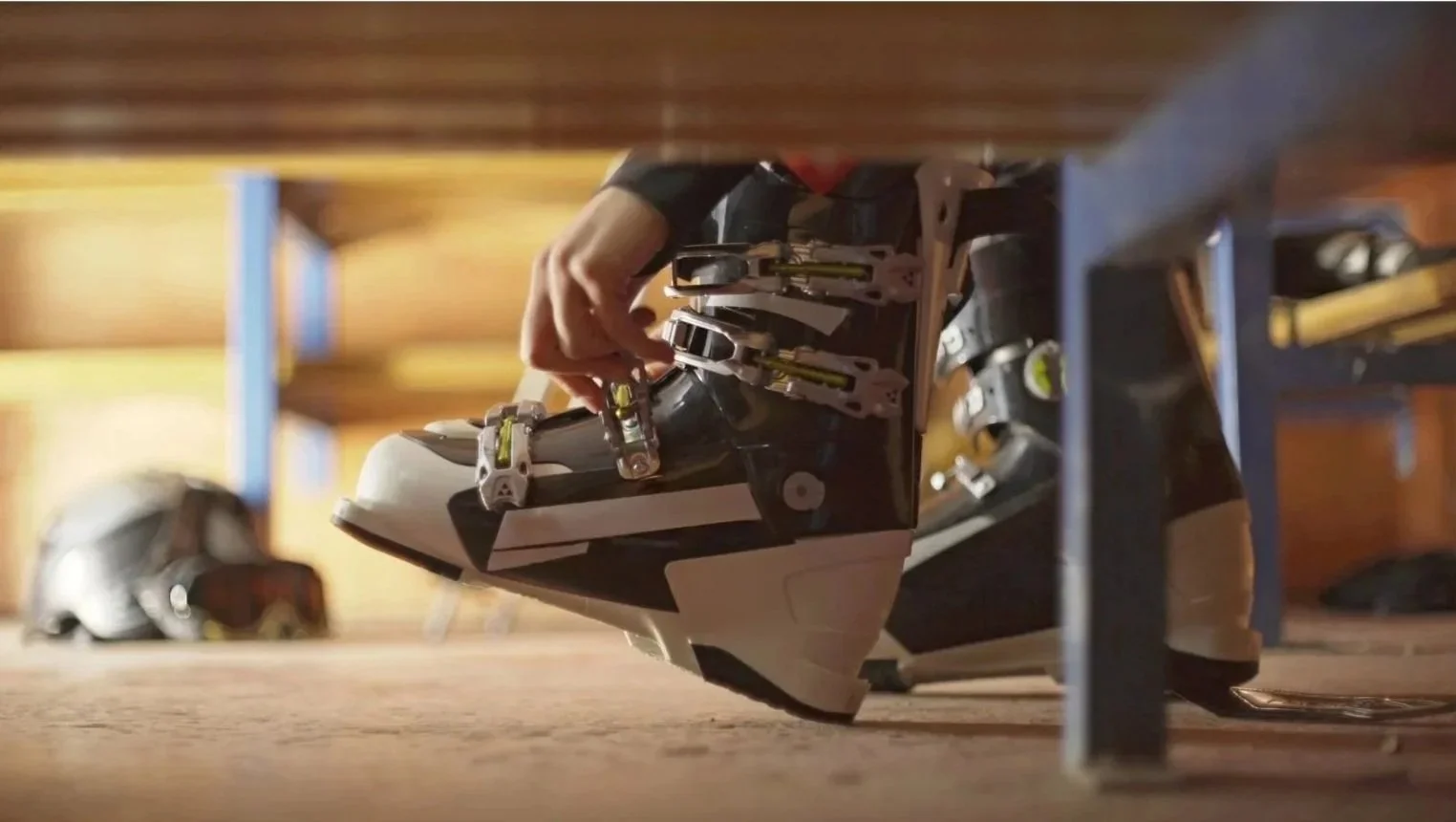 Close-up of a person adjusting ski bindings on a snow ski, with a helmet in the background, in a wooden indoor skiing facility.