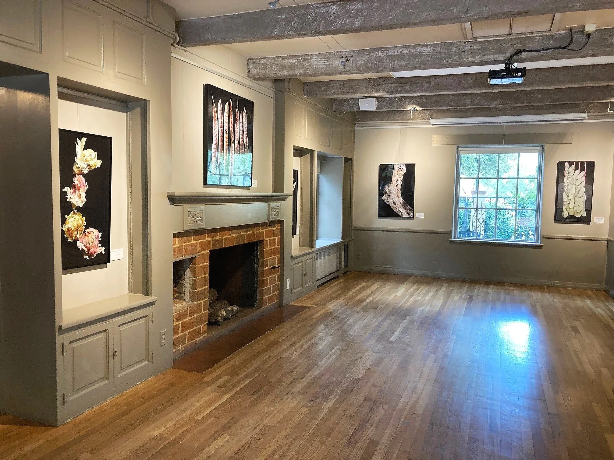 An art gallery room with light beige walls, hardwood floors, and ceiling beams. Several artworks hang on the walls, and a large window lets in natural light.