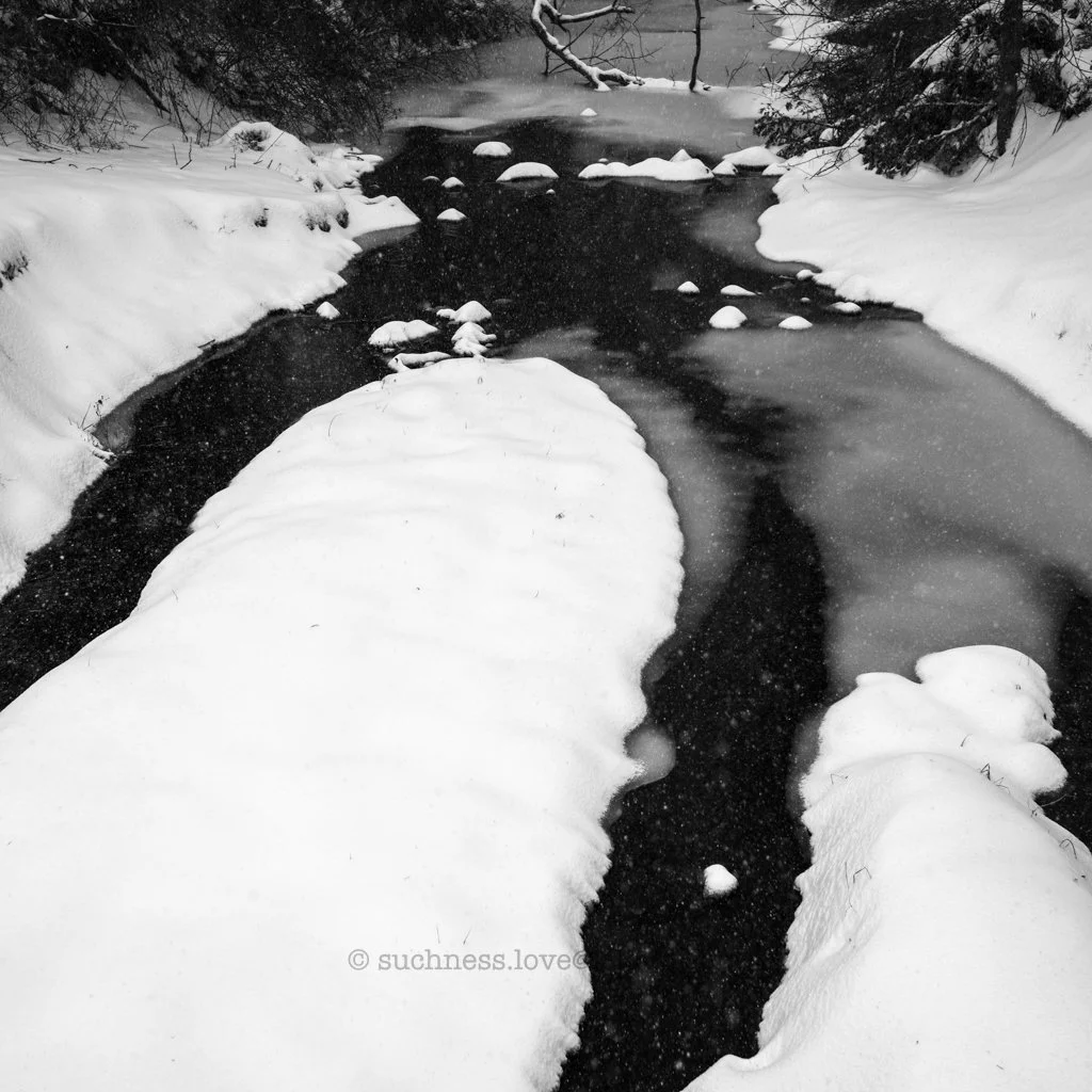 A black-and-white photo of a snow-covered riverbank with flowing water and snow-covered rocks.