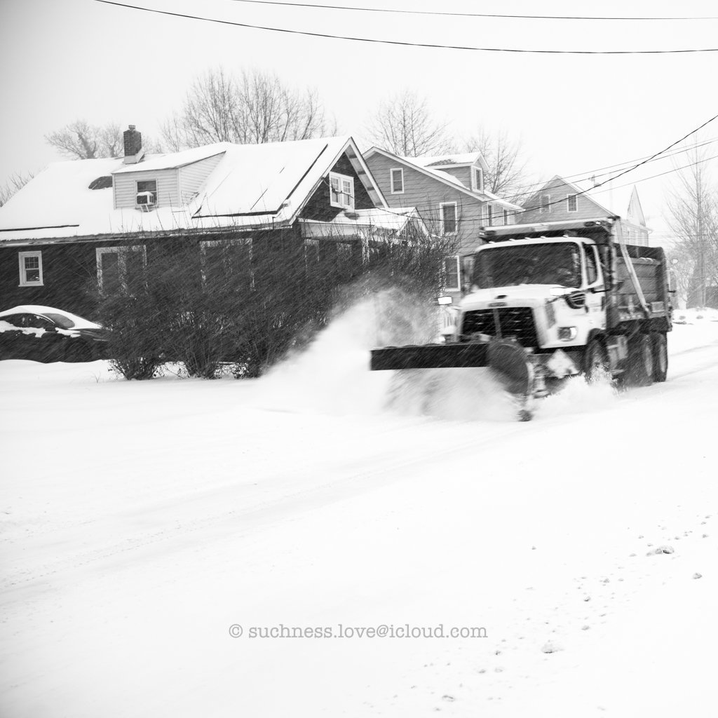 A snowplow clearing a snow-covered street in a residential neighborhood during snowy weather.