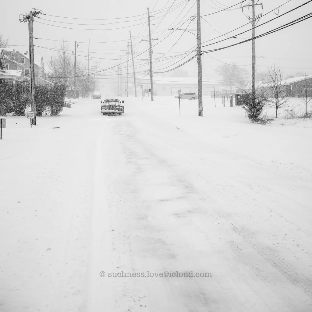 Snow-covered street with cars, power lines, and houses, during a snowstorm in a residential neighborhood.