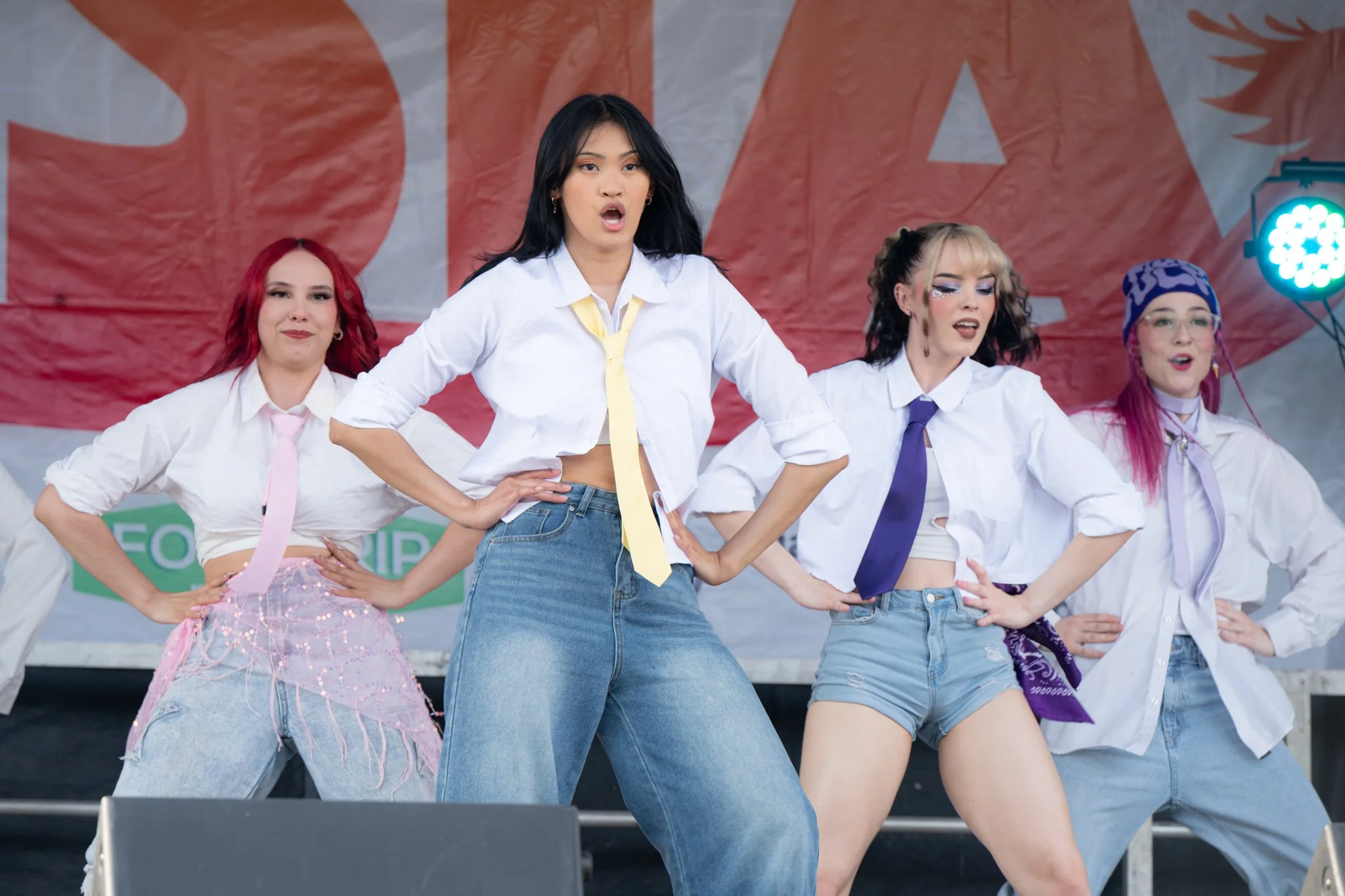 Group of five women performing on an outdoor stage, dressed in colorful and stylish clothing, with a red banner in the background.