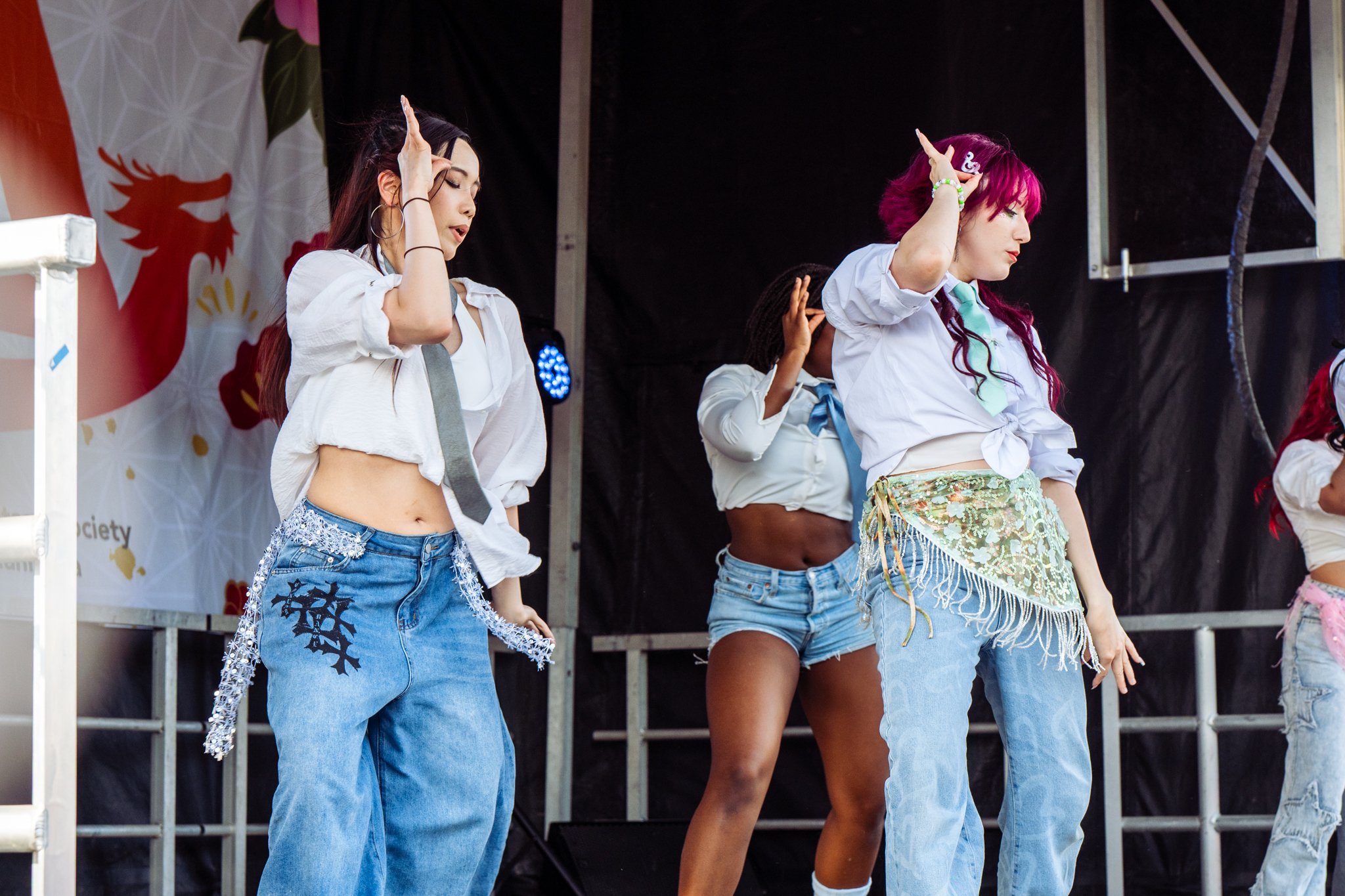 Three young women performing a coordinated dance on an outdoor stage during daytime, dressed in colorful, casual clothing with some traditional elements.