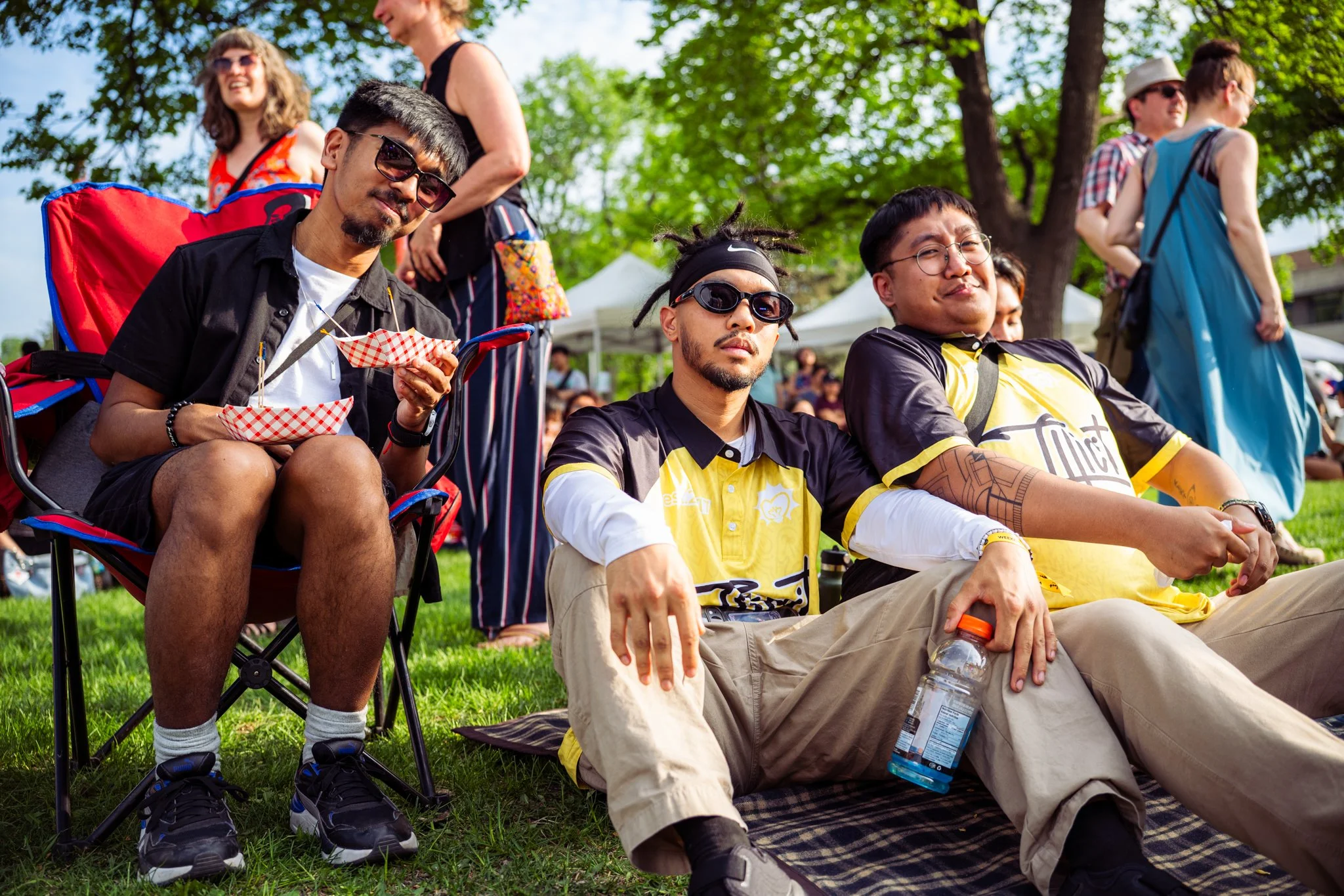 Group of people sitting and standing outdoors on a sunny day, with green trees in the background. Some are seated on chairs and blankets, enjoying the event, with one person holding a water bottle and another eating snacks.