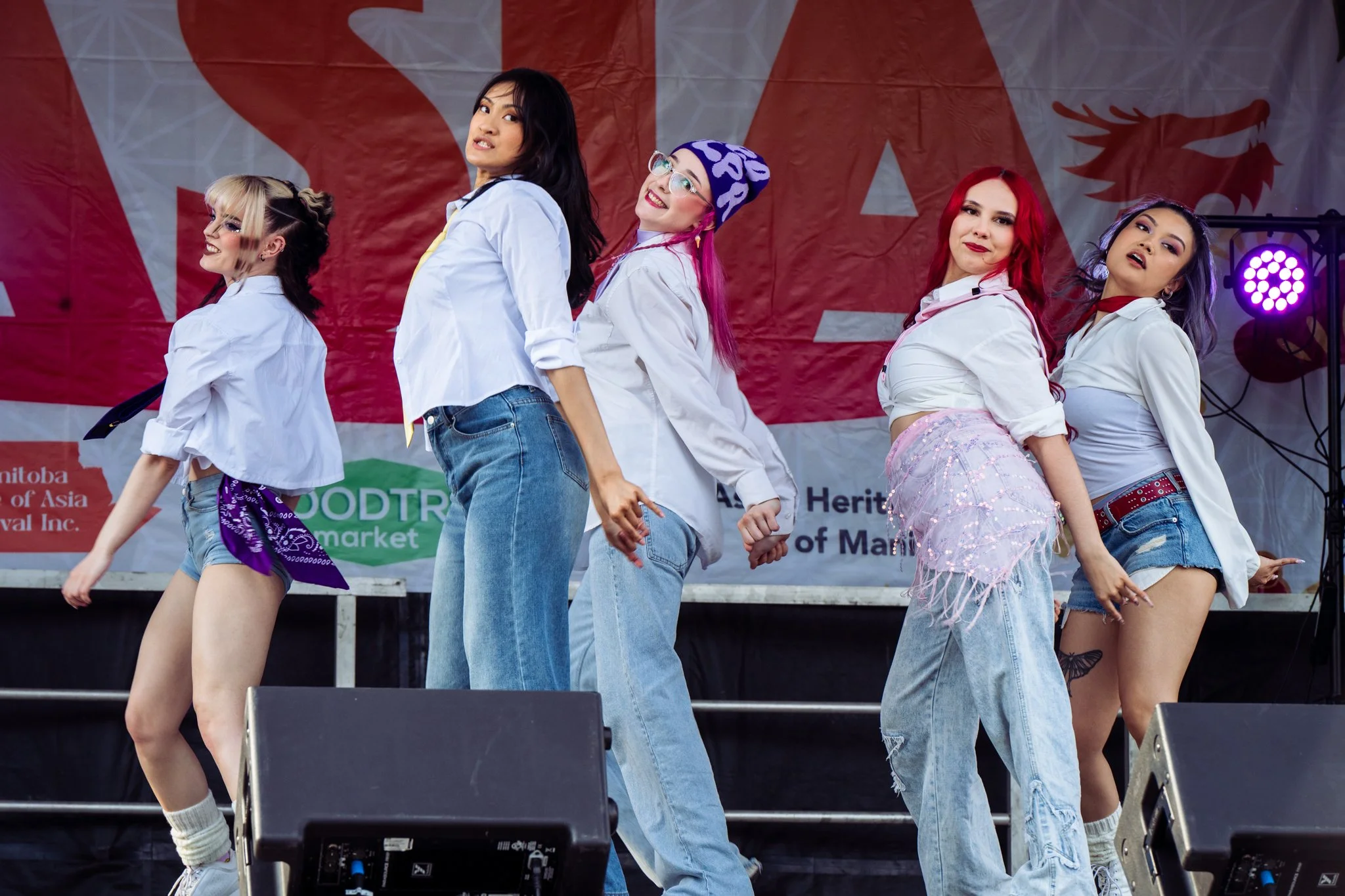 Six women performing a dance on stage with a red backdrop featuring a lion and text, dressed in casual attire including jeans and white shirts, some with accessories like a purple bandana and a beanie.