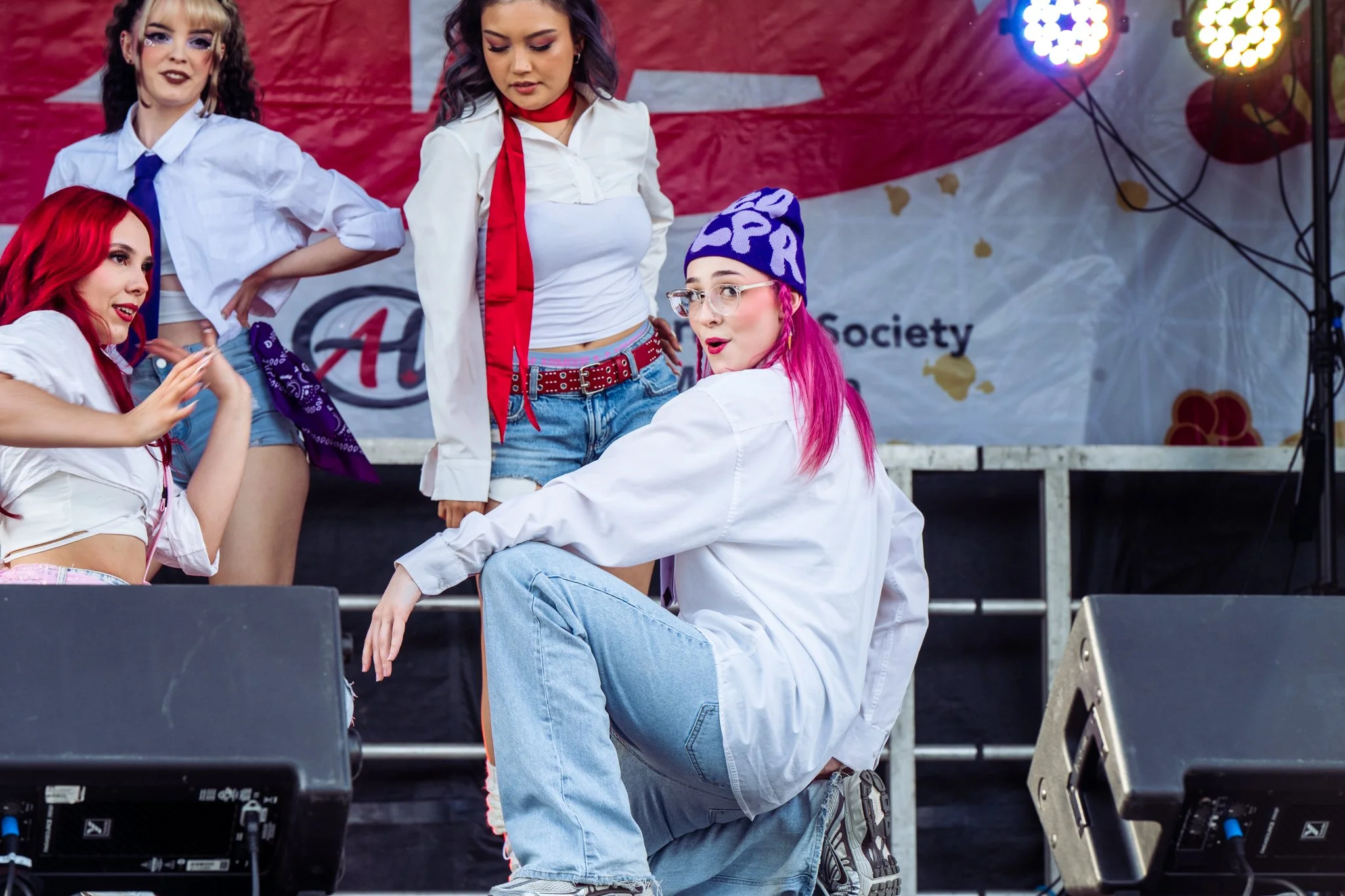 Group of young women with colorful hair and stylish outfits on stage during a performance or event, with stage lights in the background.