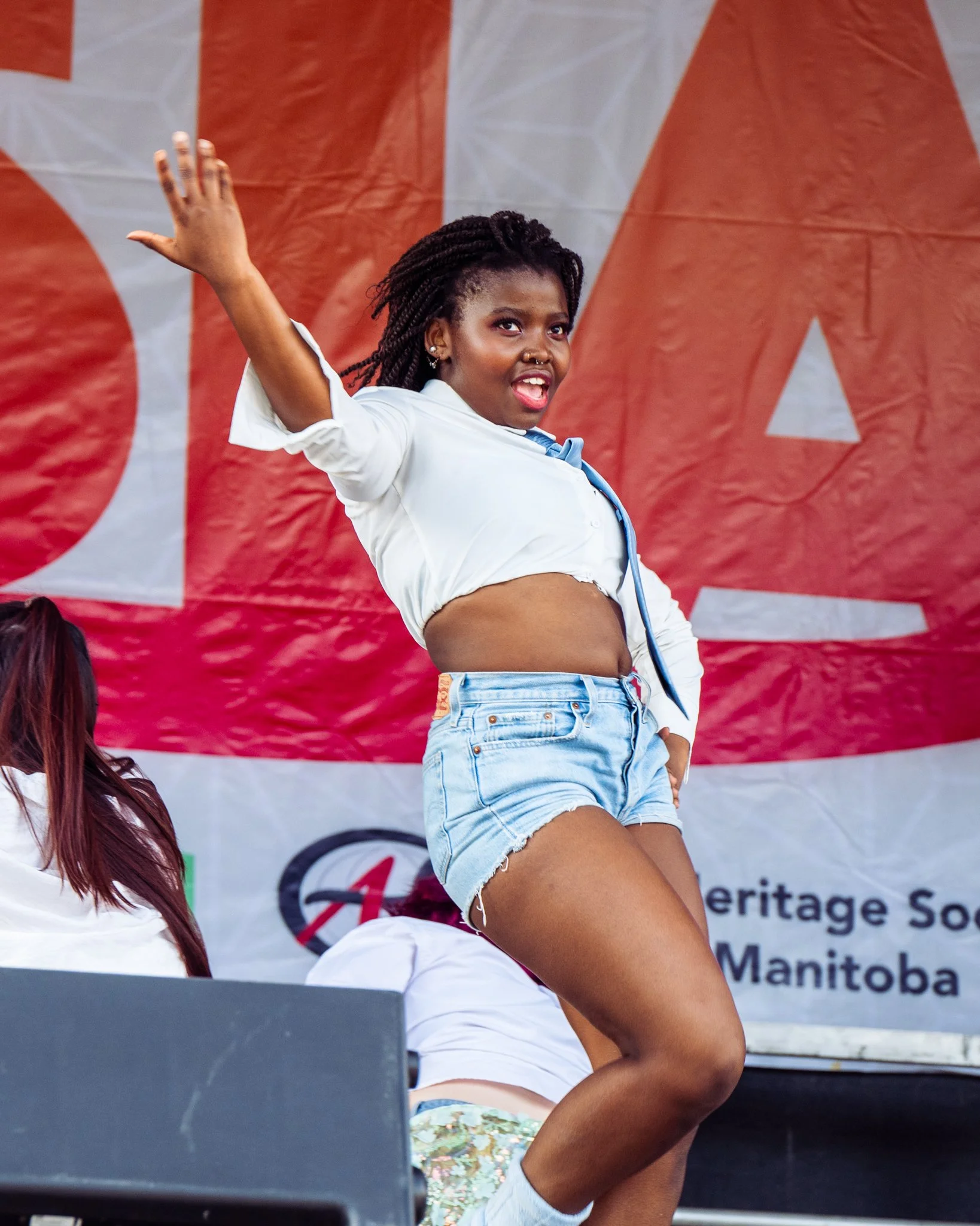 A woman with braided hair in a white shirt and denim shorts, dancing or performing on stage with a red and white backdrop.