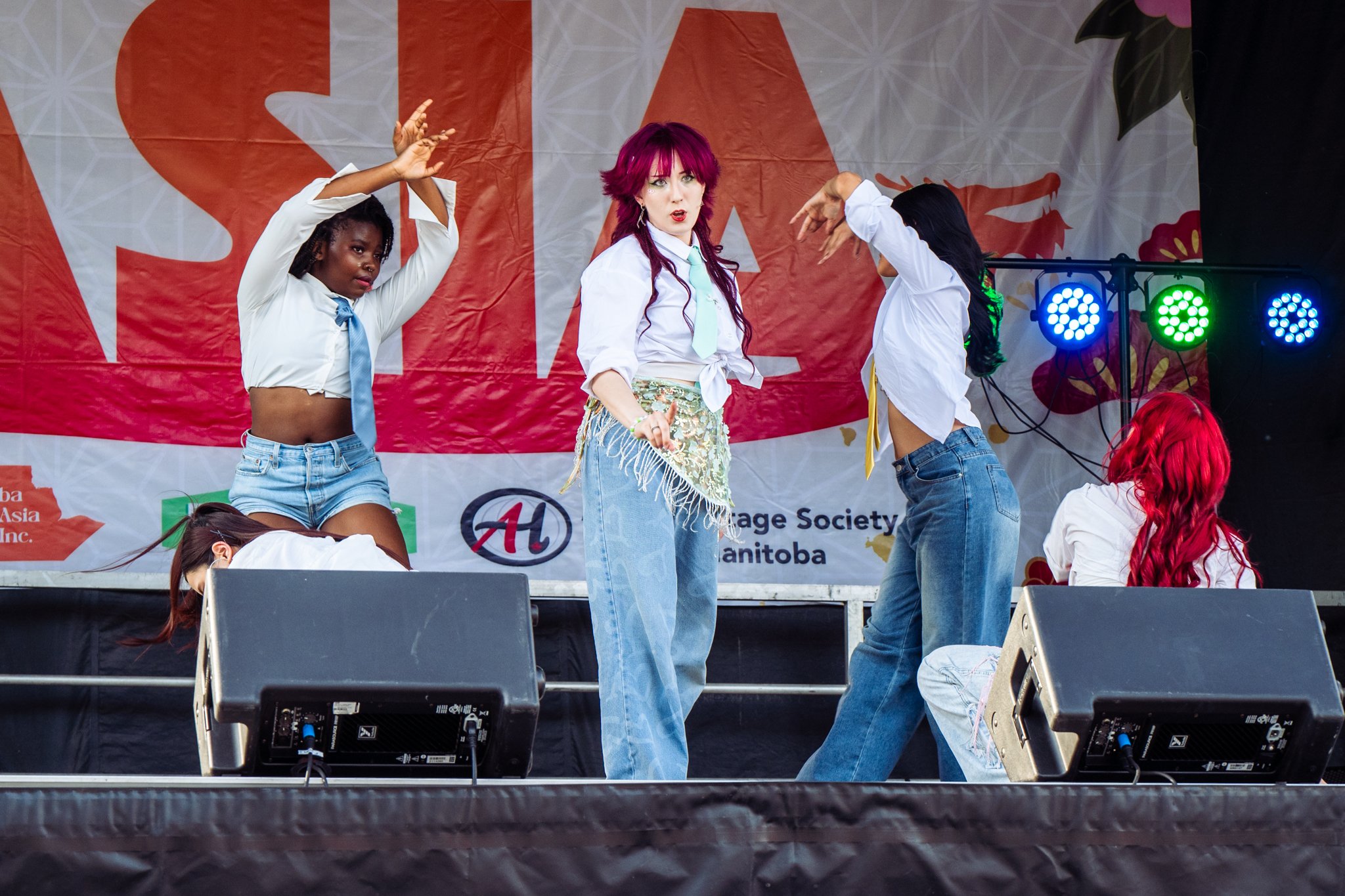Group of three women performing on stage with a red and white banner in the background. The woman in the center has bright pink hair, white shirt with rolled-up sleeves, a light blue tie, and a decorative skirt. The woman on the left has dark braided