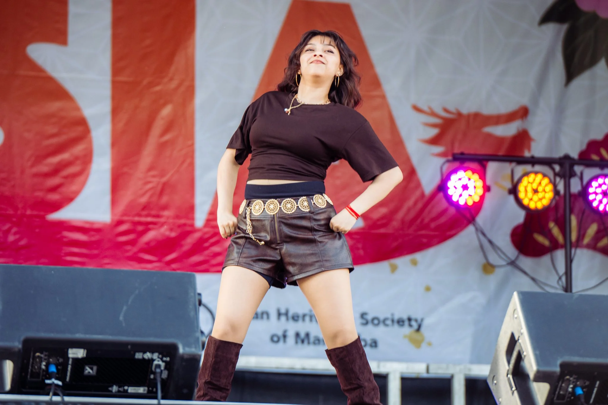 Young woman performing on stage, wearing a black top, leather shorts with a decorative belt, and knee-high boots, with colorful stage lights in the background.
