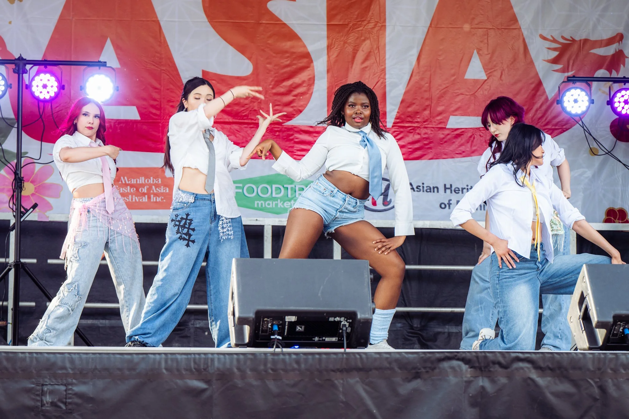 Five young women dancing on a stage during a performance at an outdoor festival, with colorful backdrop and stage lights.