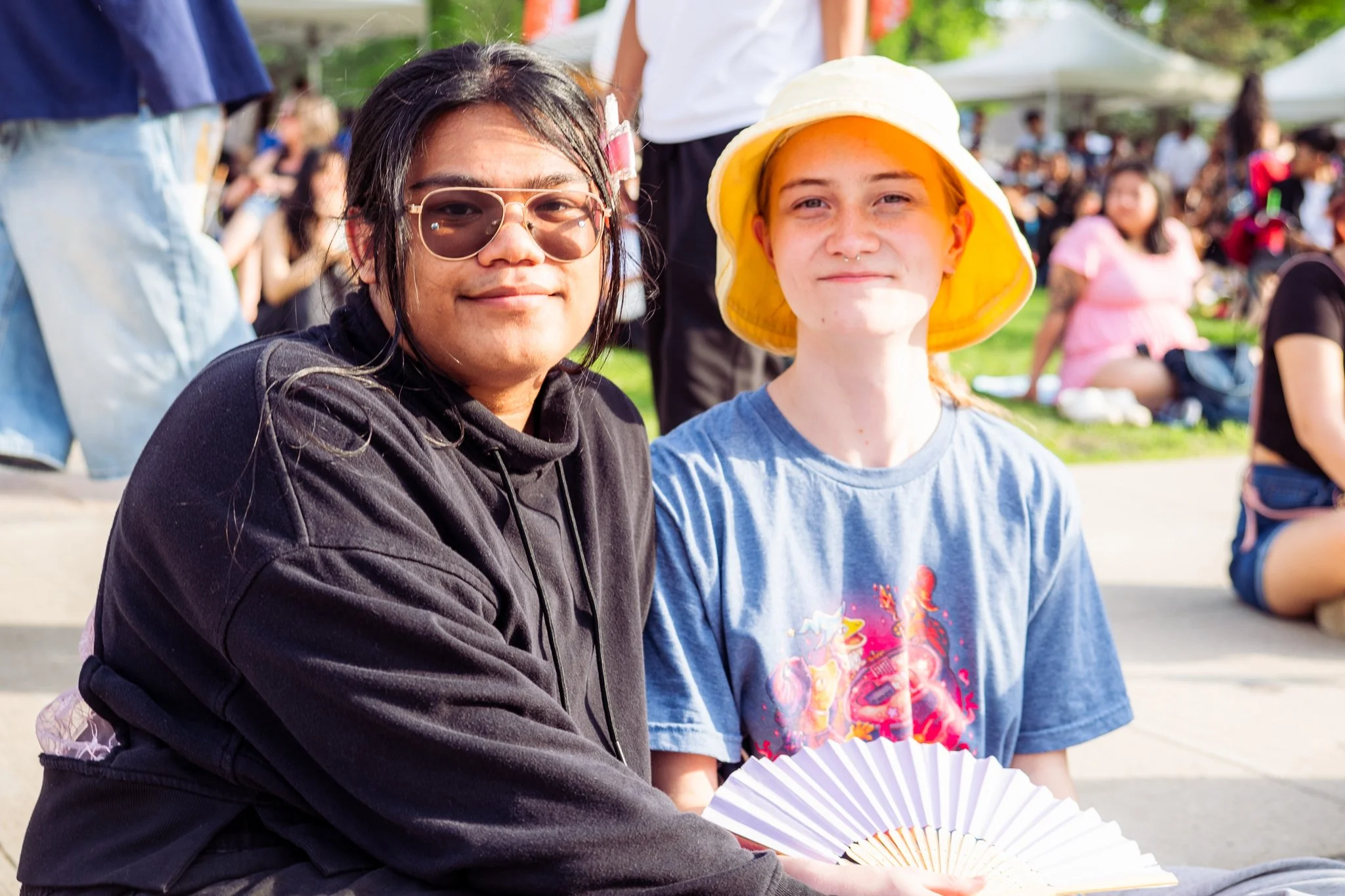 Two young people sitting outdoors at a public event or festival, with a crowd in the background. The person on the left has dark hair, glasses, and is wearing a black hoodie. The person on the right has red hair, a yellow sun hat, and is holding a pa