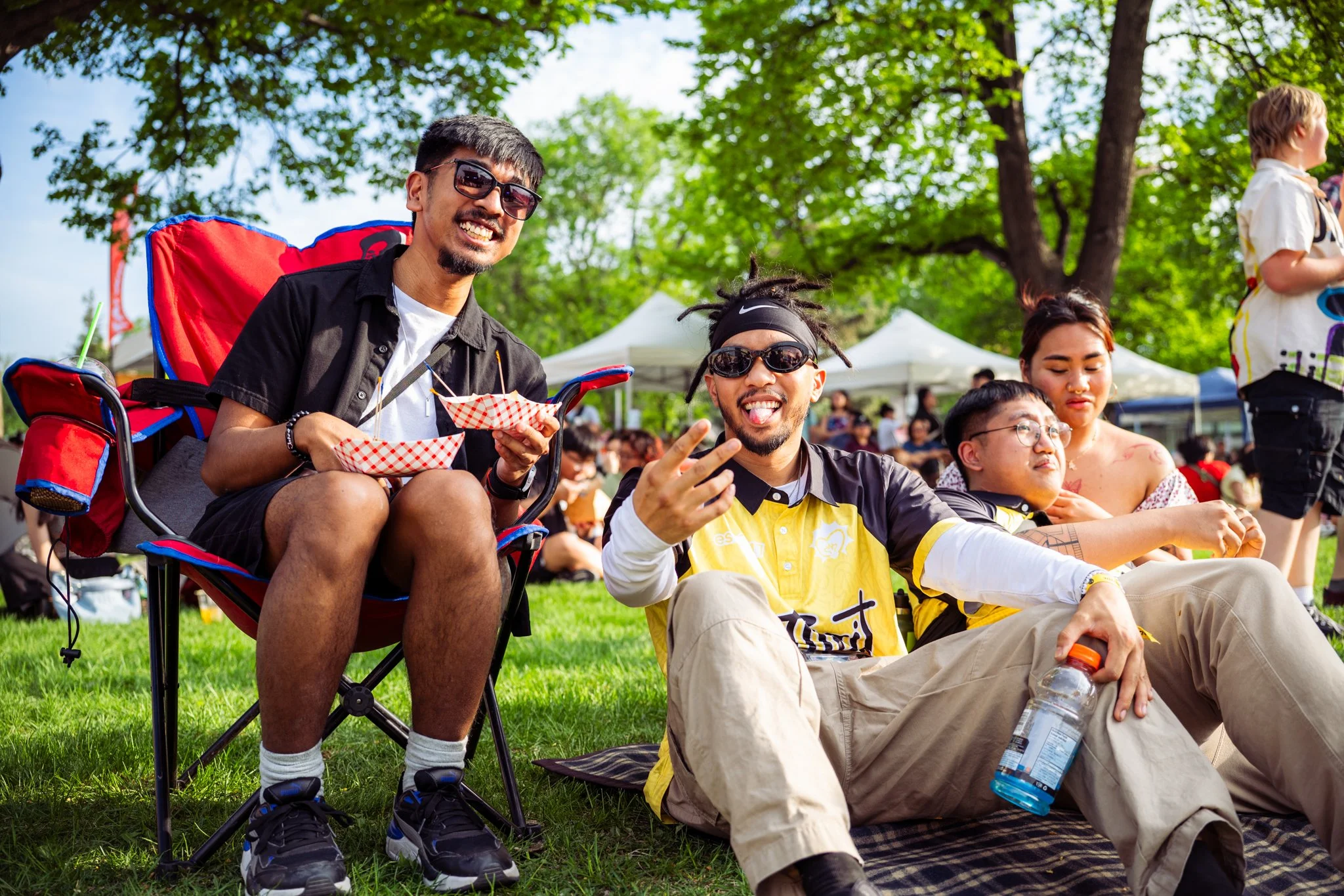 A group of diverse people sitting on the grass at an outdoor event, smiling and enjoying food and drinks under green trees on a sunny day.