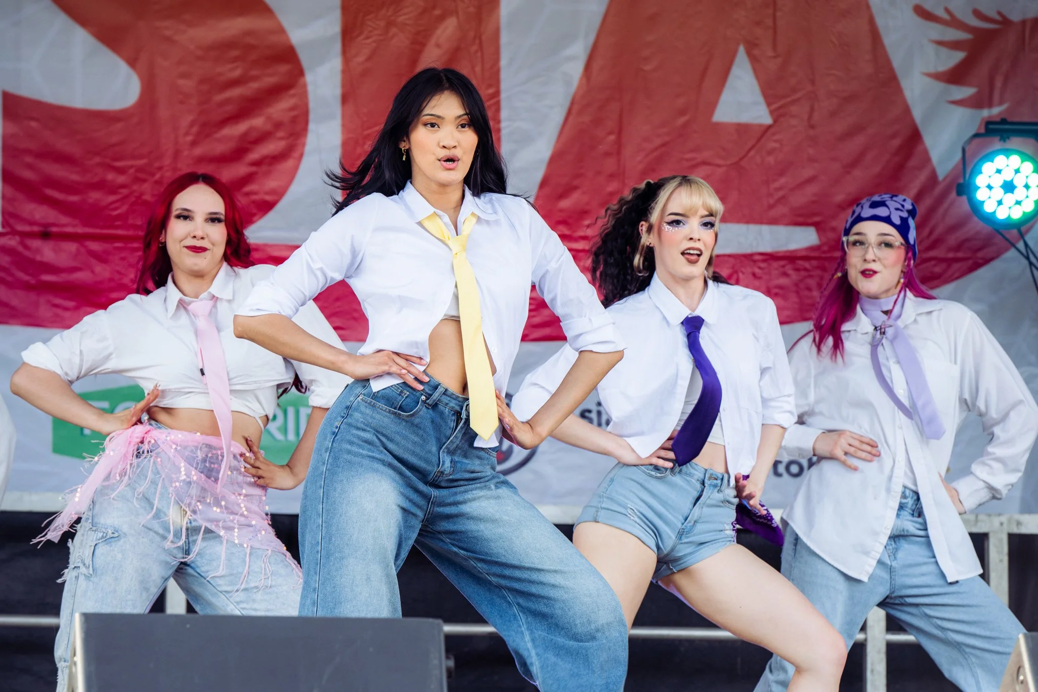 A group of five women performing a dance on stage at an outdoor event, with a large red and white banner in the background. The women are dressed in colorful, casual outfits with some wearing ties and accessories.