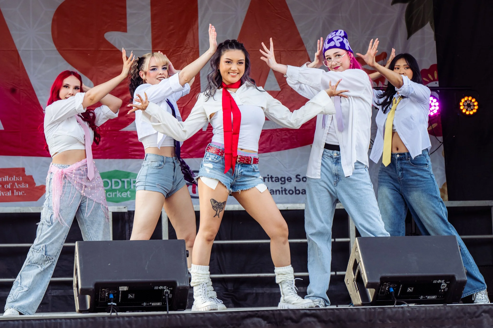 A group of six young women performing a dance on stage. They are wearing casual outfits including jeans, skirts, and colorful accessories. The stage has a background with red, white, and green colors and some stage lights visible.