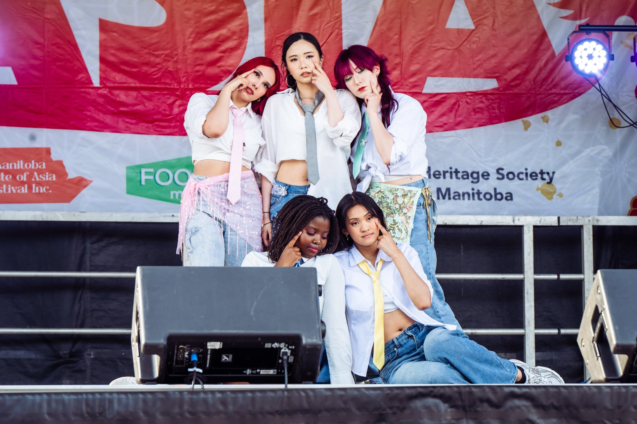 Five young women on an outdoor stage posing with fingers on their faces, with a red and white banner behind them and stage lights on the sides.