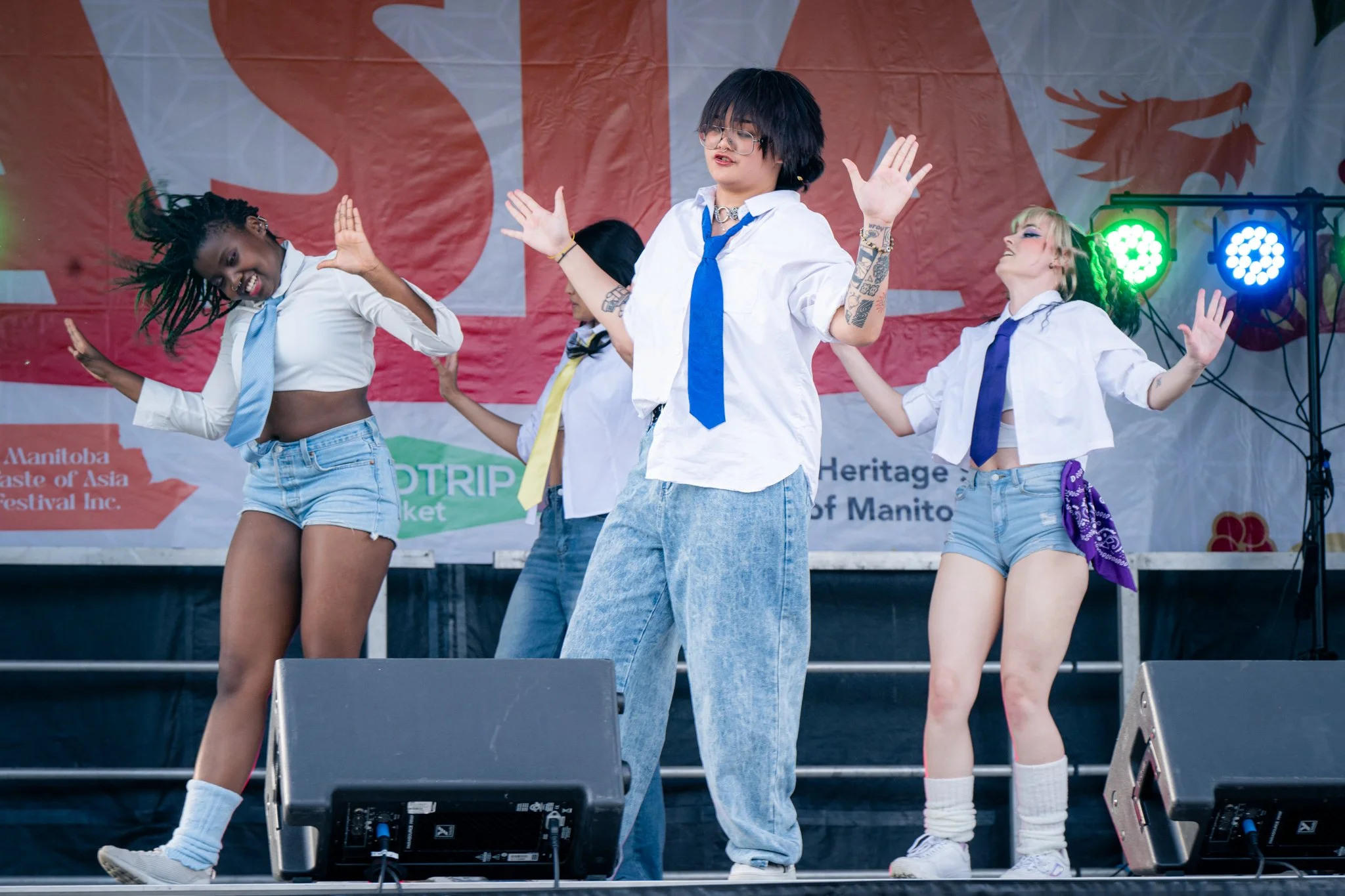 Group of diverse young women dancing on stage at a festival, with a large red and orange banner in the background and colorful stage lights.
