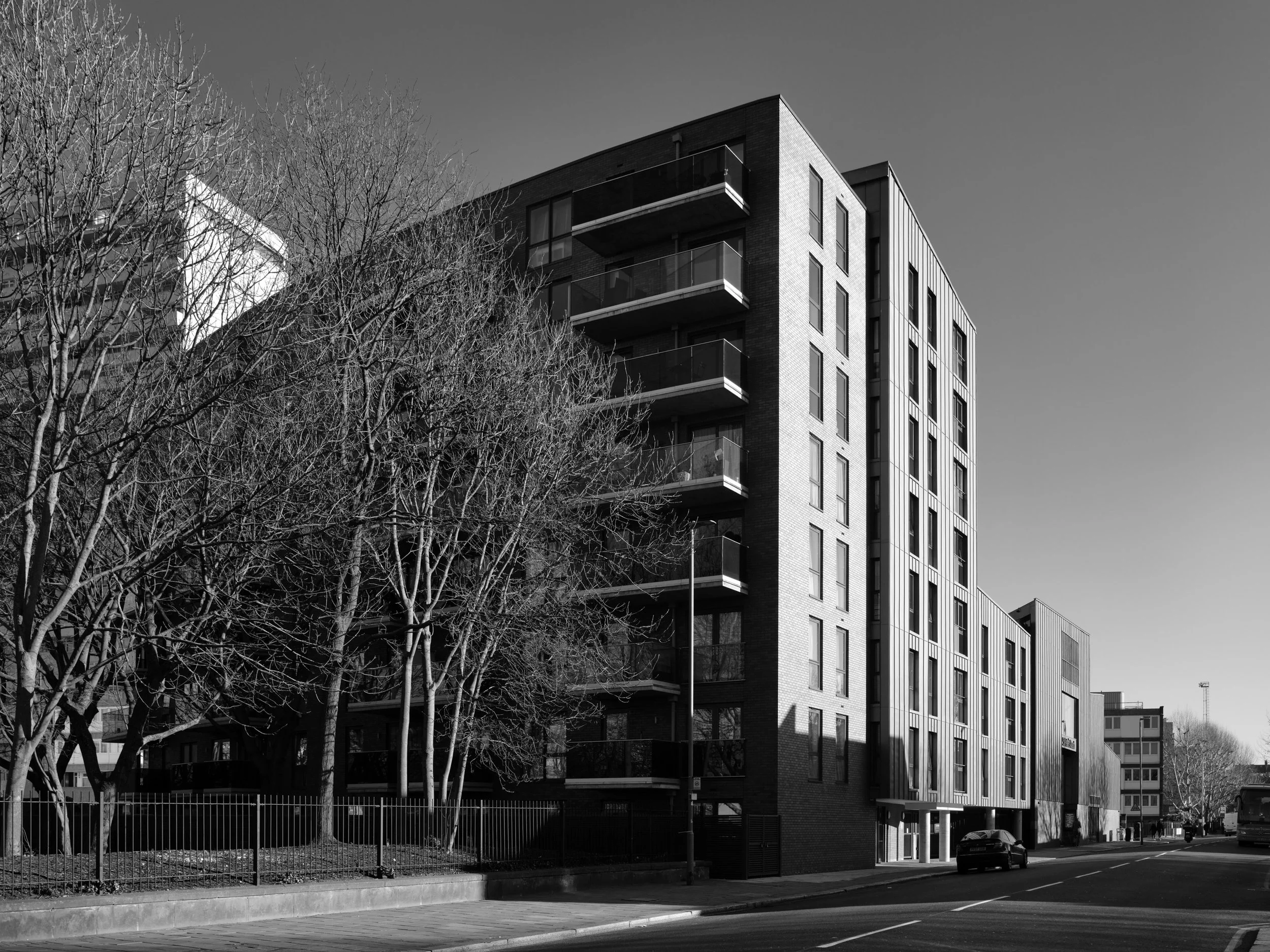 Multiple people walking and biking on a paved walkway in front of modern multi-story apartment buildings surrounded by green trees on a sunny day.