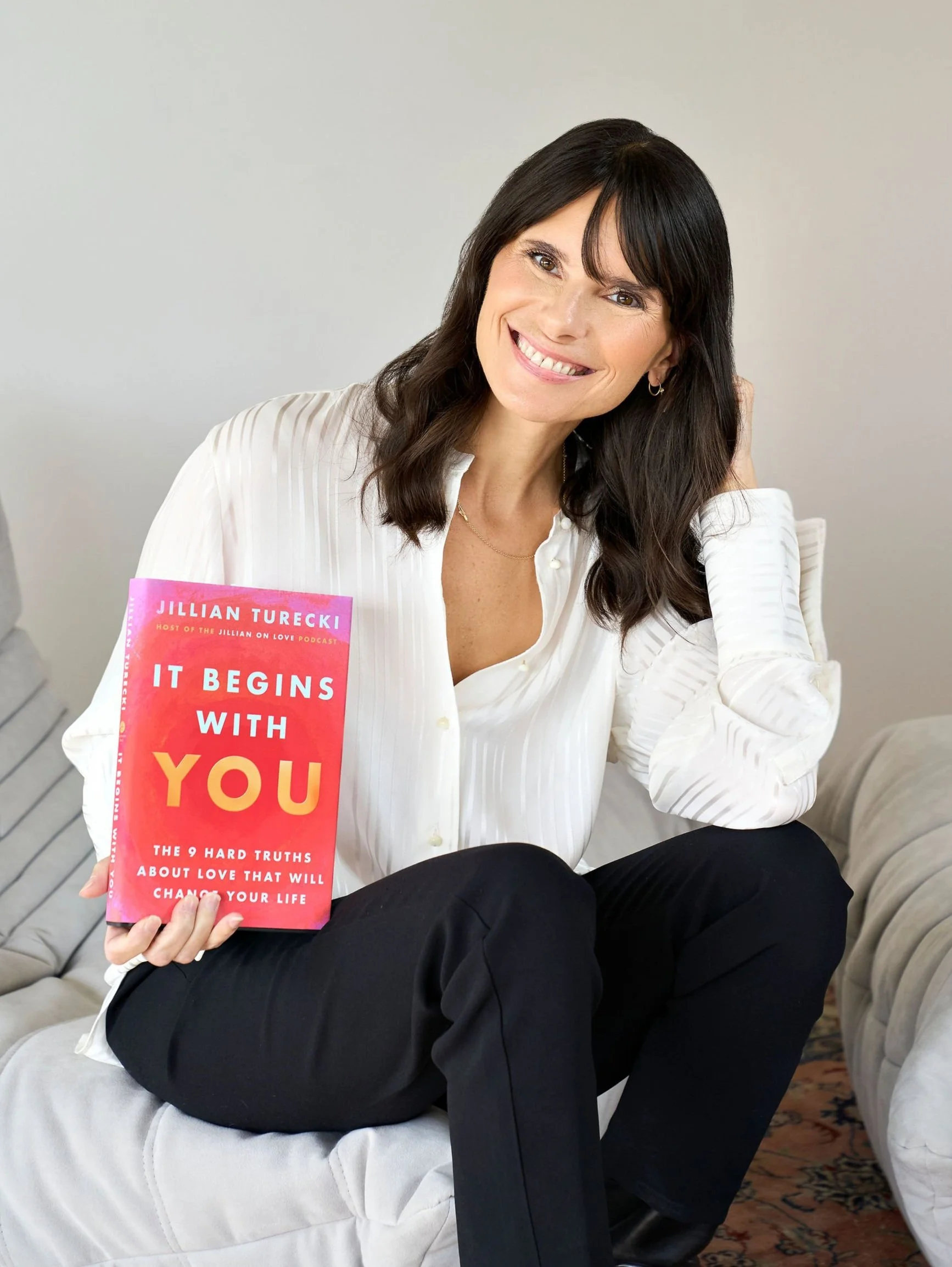 Jillian Turecki in a white blouse sitting on a sofa, holding a pink and orange book titled "It Begins With You" by Jillian Turecki, smiling at the camera.