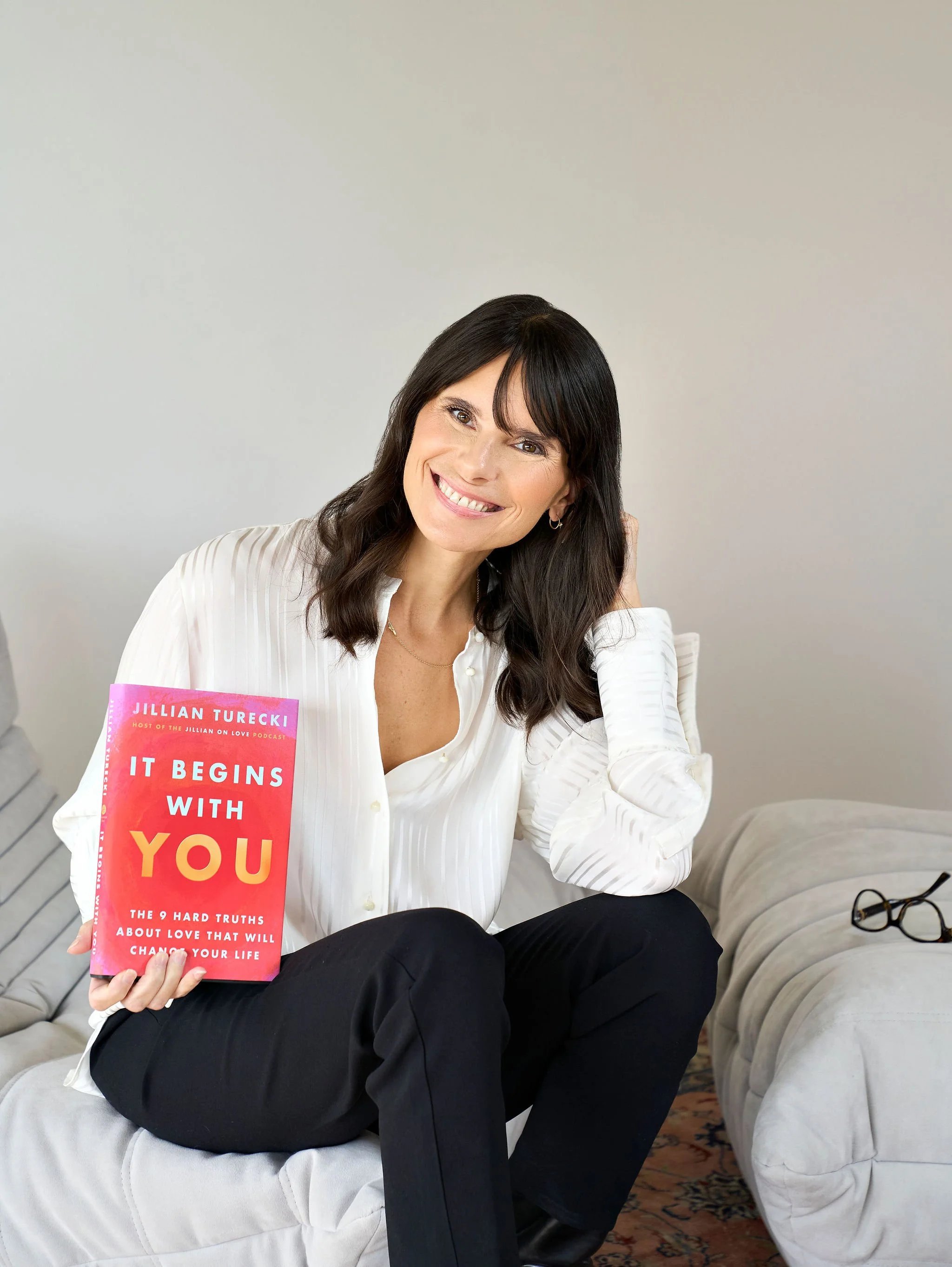 Jillian Turecki wearing a white blouse and black pants sitting on a white couch holding a book cover titled 'It Begins With You' by Jillian Turecki, with subtitle about hard truths on love, featuring a pink and orange watercolor background.